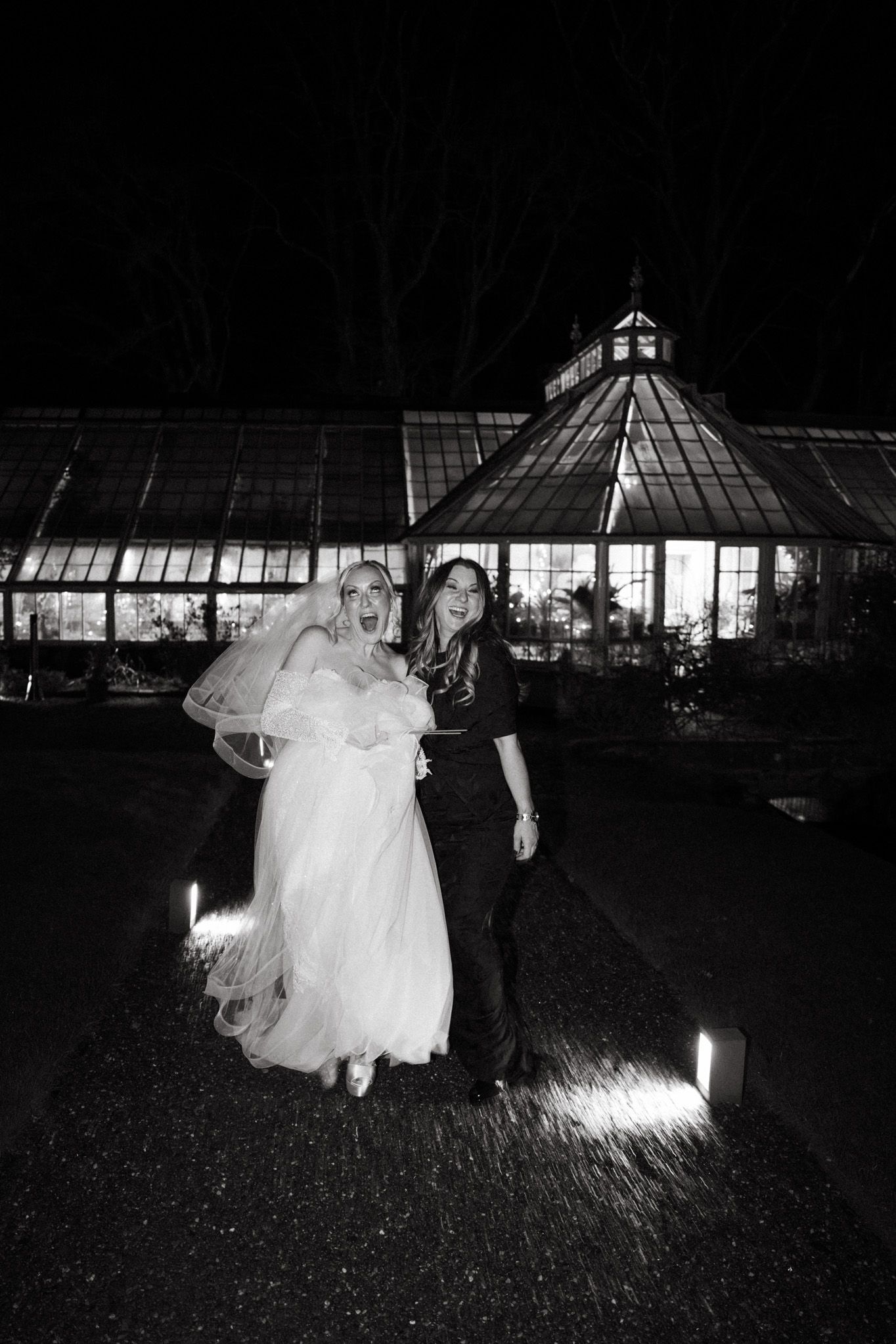Atmospheric black and white photo of wedding celebration outside illuminated greenhouse.