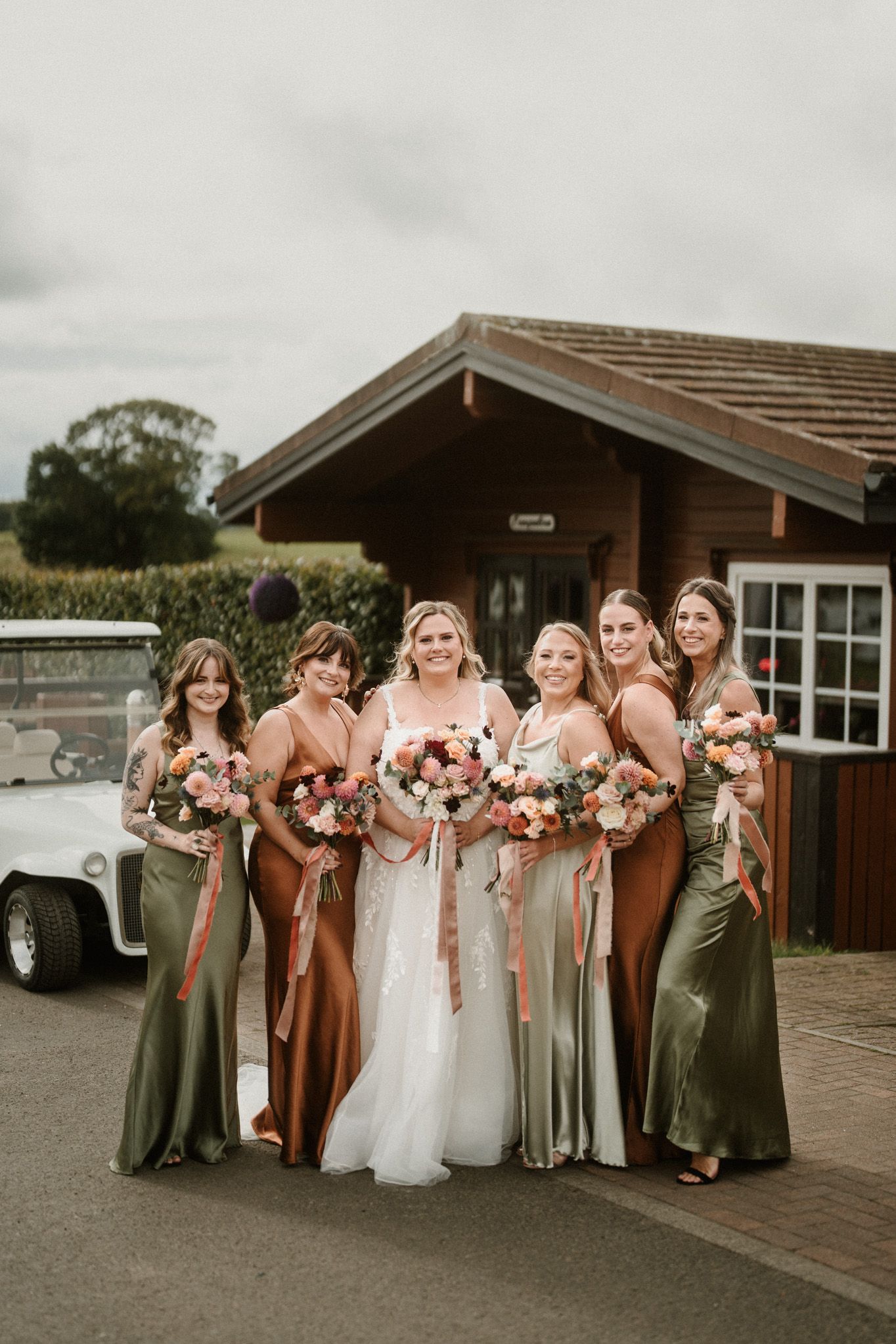 Wedding party in olive green and rust colored dresses pose together with bouquets outside a rustic wooden venue.
