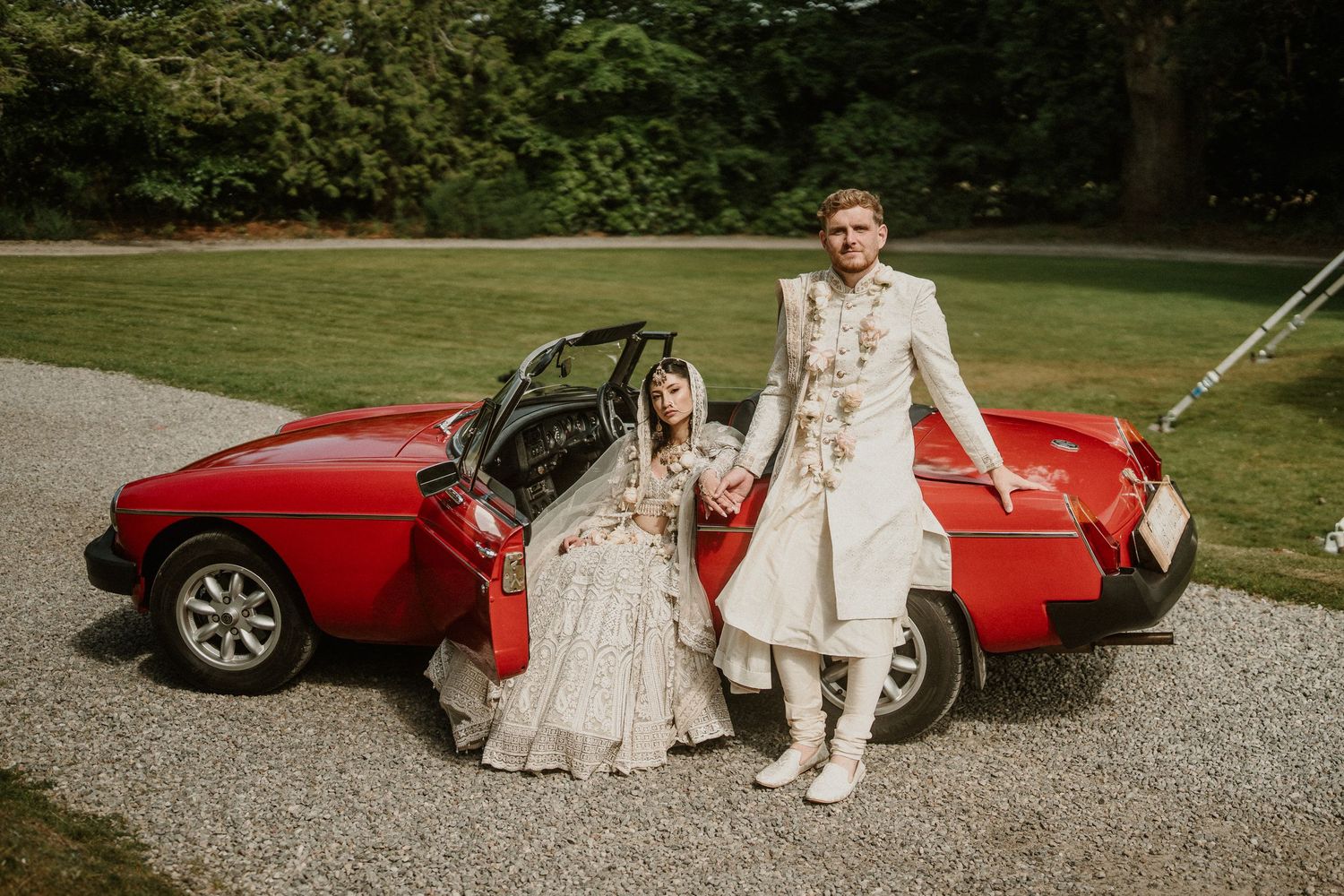 Wedding couple relaxing by vintage red sports car on gravel driveway.