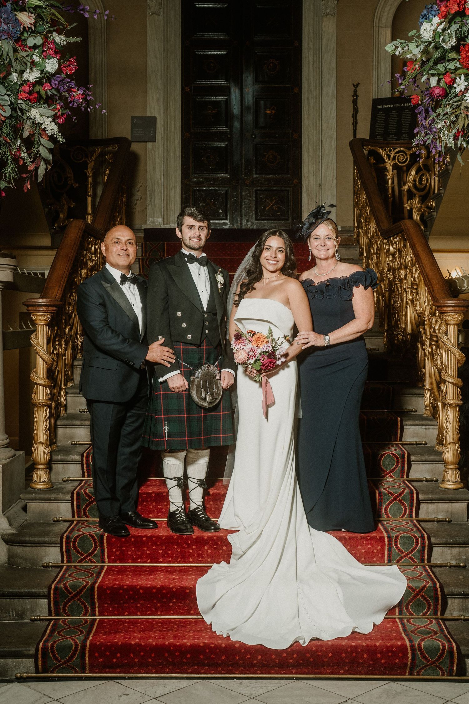 Wedding party poses on grand staircase with red carpet and ornate gold railings.