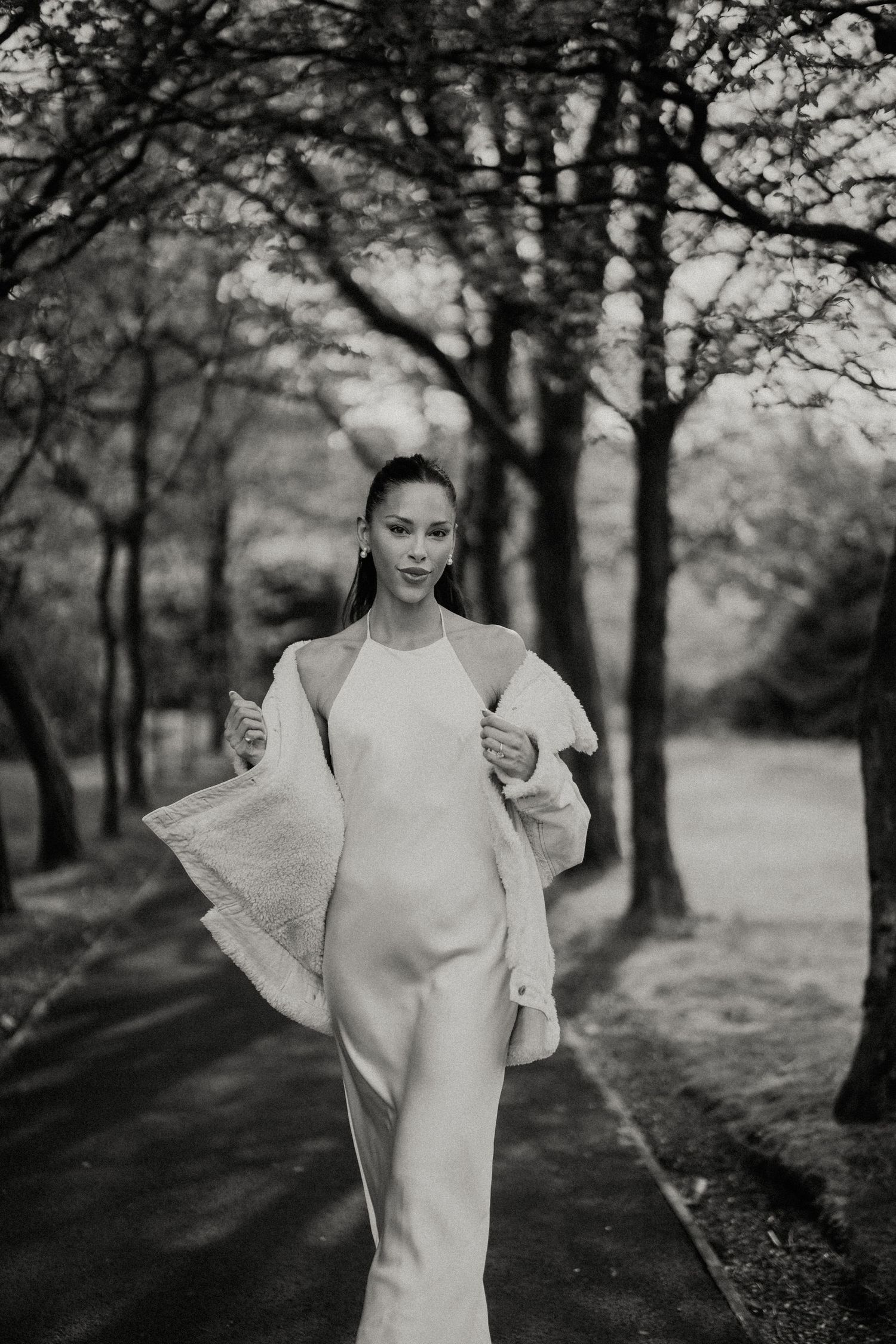 An elegant figure poses in a white dress on a tree-lined path in black and white.