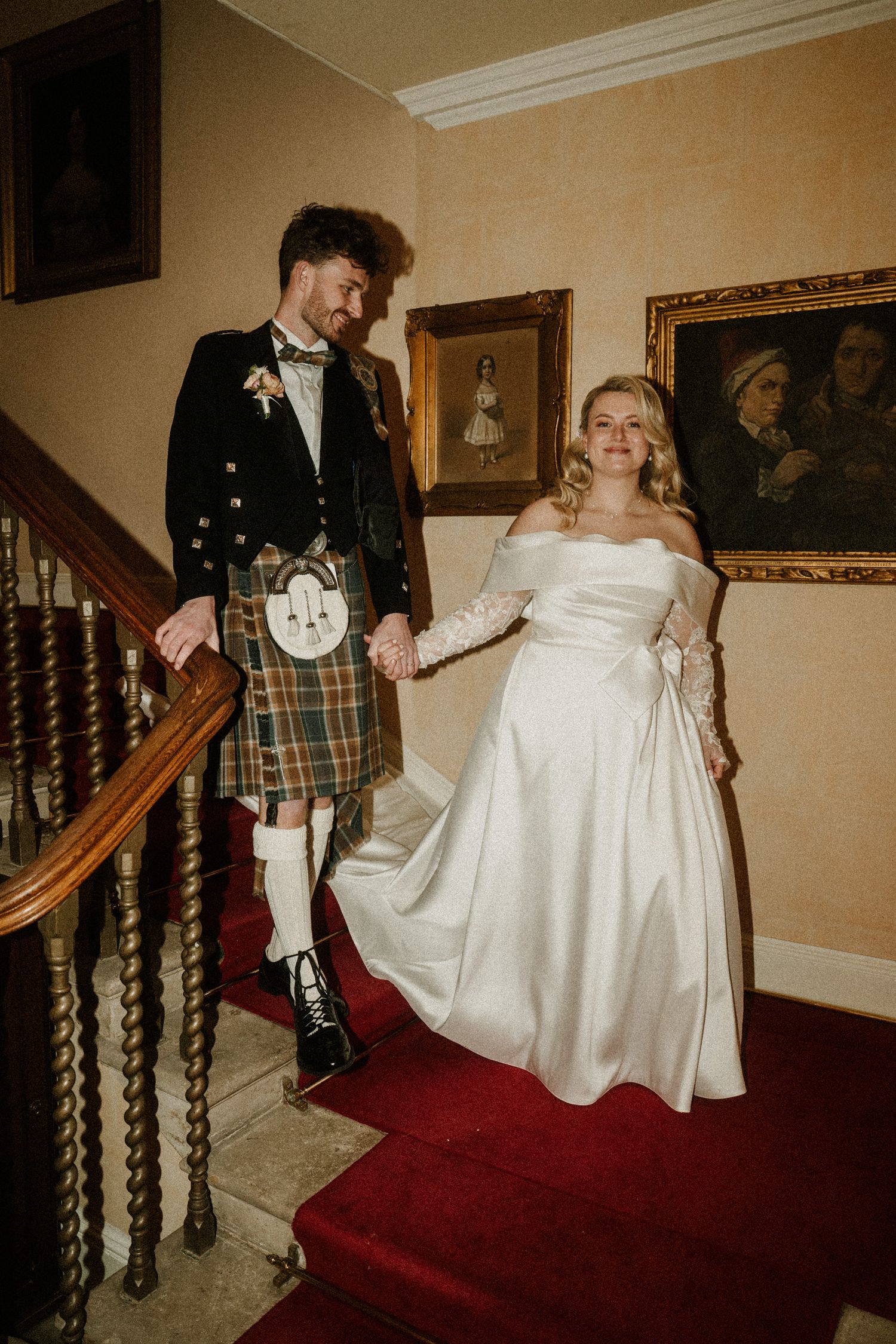 Couple in Scottish wedding attire posing on red-carpeted stairs in historic building.
