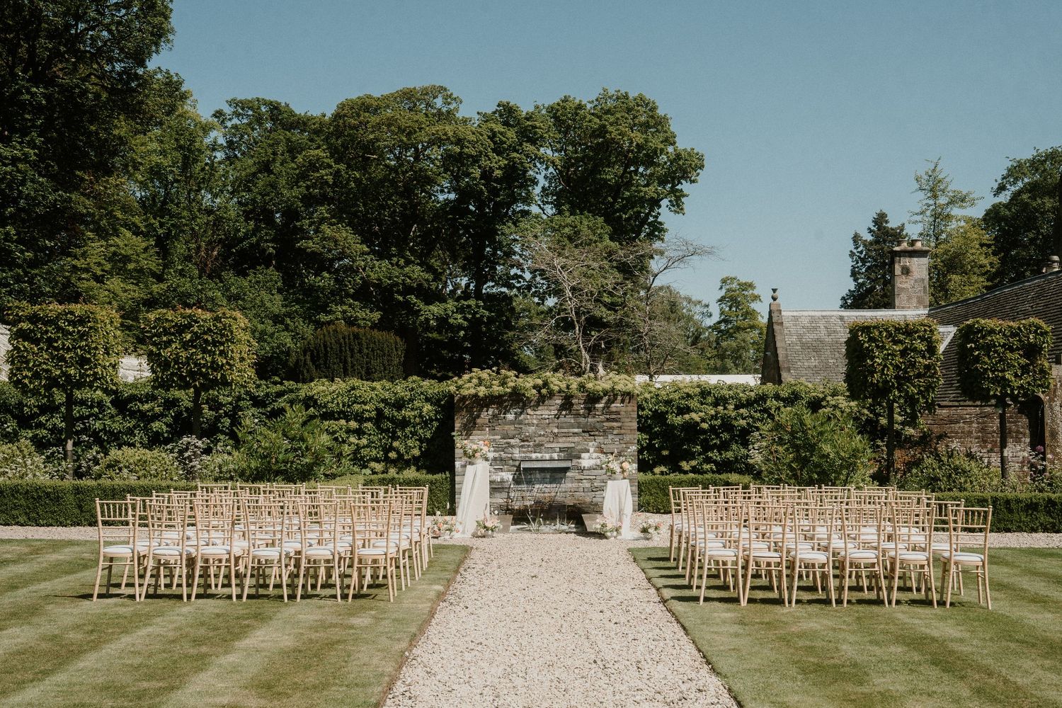 Outdoor wedding ceremony setup with rows of wooden chairs on a gravel path leading to a stone wall backdrop.