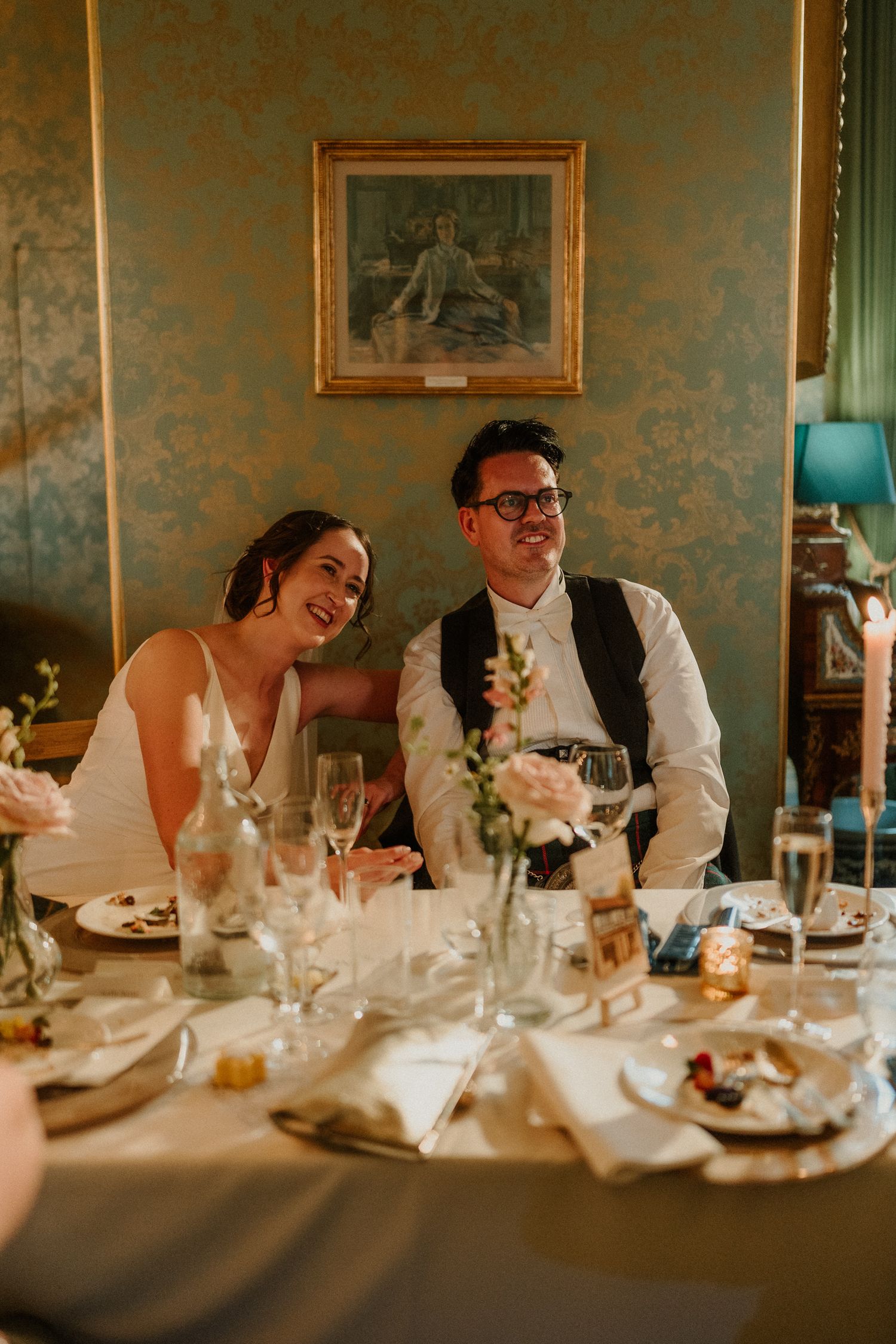 A smiling bride and groom sit together at a elegantly set wedding reception table in a vintage-styled room.