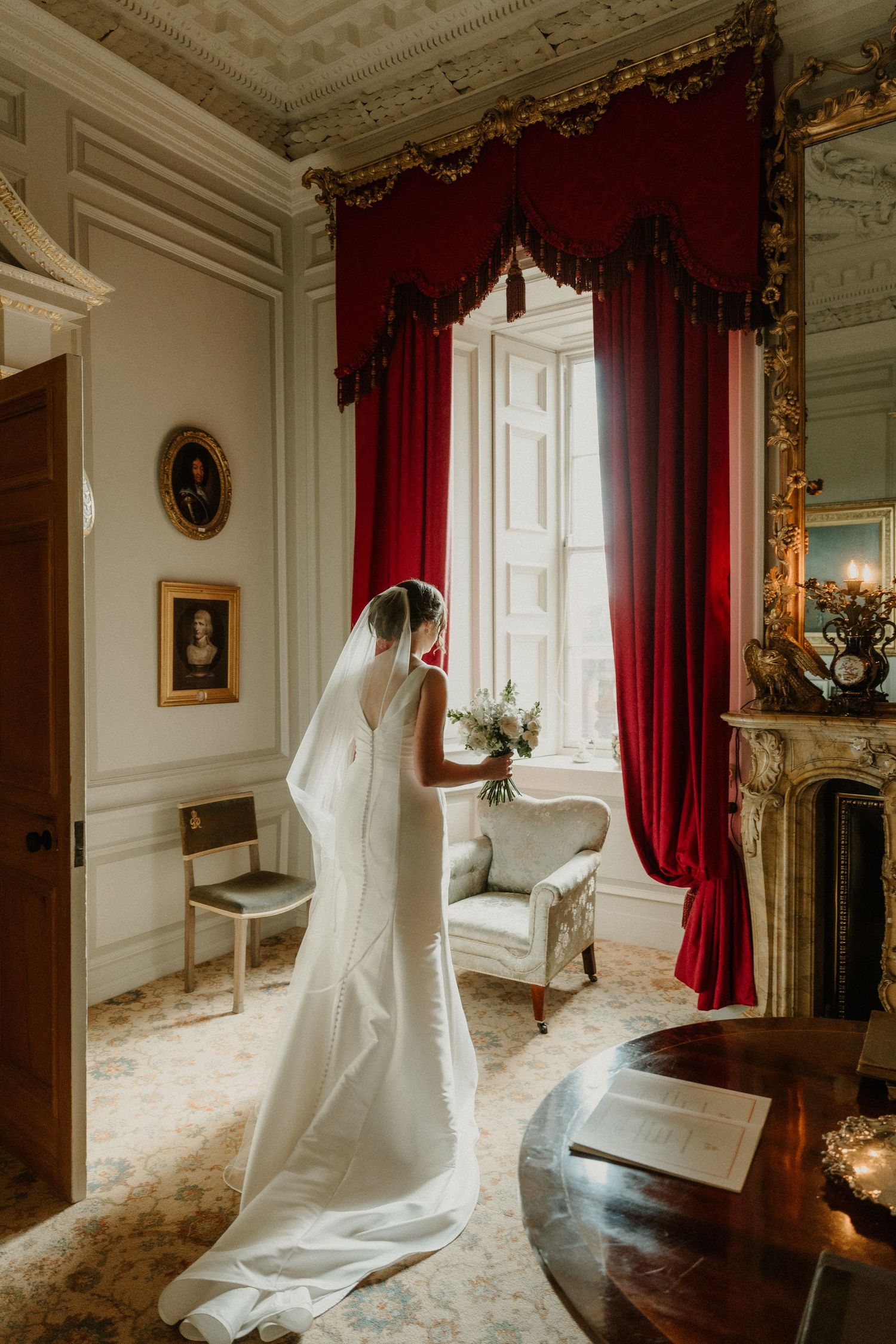 Bride in white gown and veil holds bouquet by ornate window with red drapes in elegant manor room.