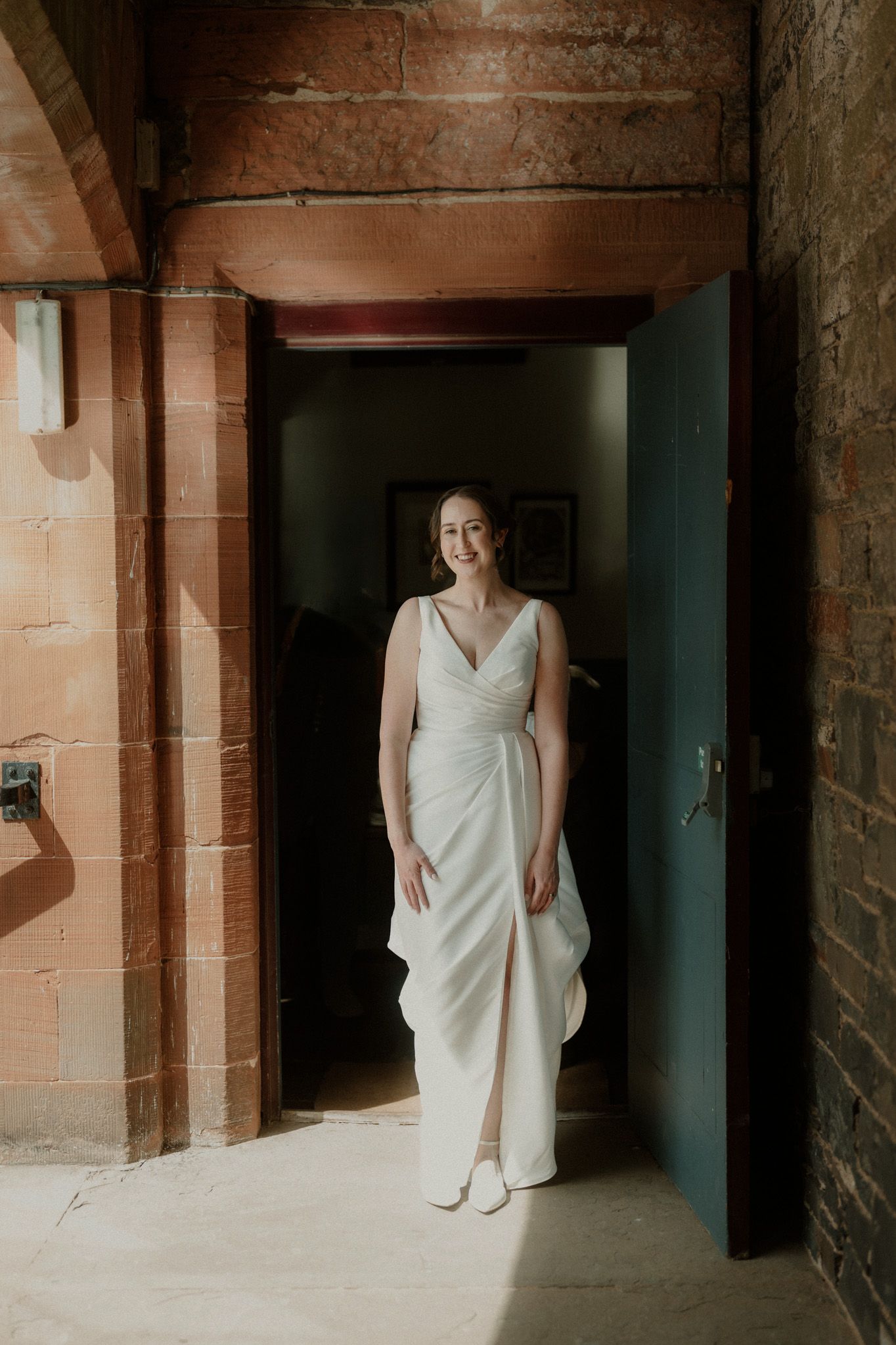 Smiling bride in an elegant white wrap-style wedding dress stands in a sunlit red sandstone doorway with an open teal door.