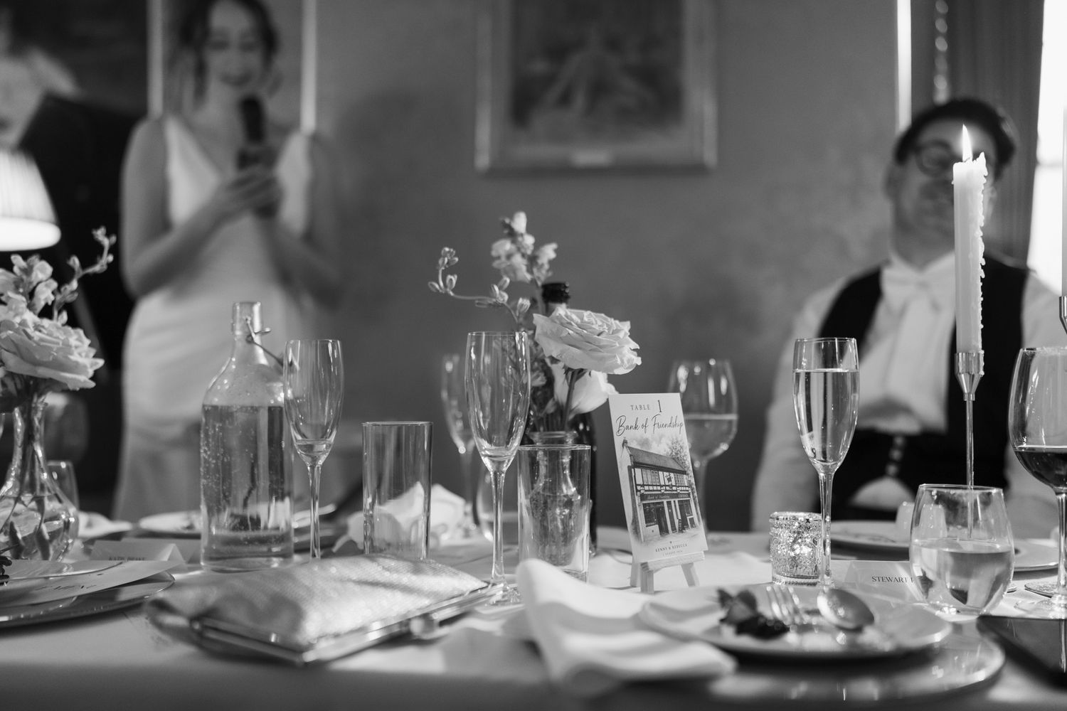 Black and white wedding reception table with candles, wine glasses, flowers and plates as guests in formal attire sit in the background.