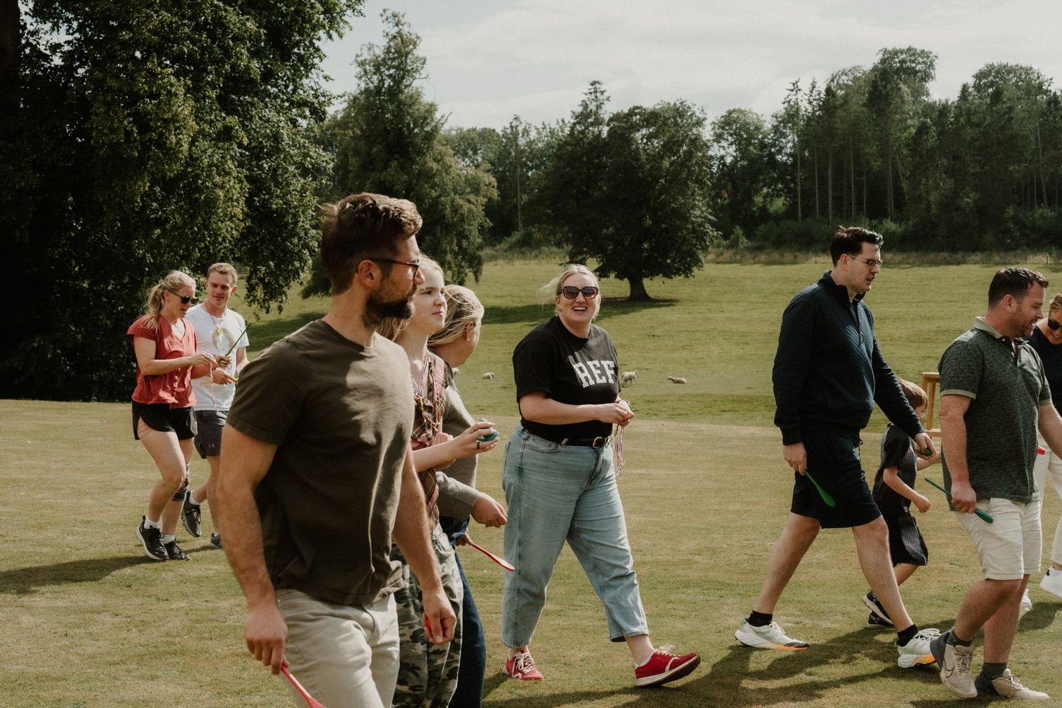 Group of people in casual clothes walking together across a sunny grassy field with tall green trees in the background.
