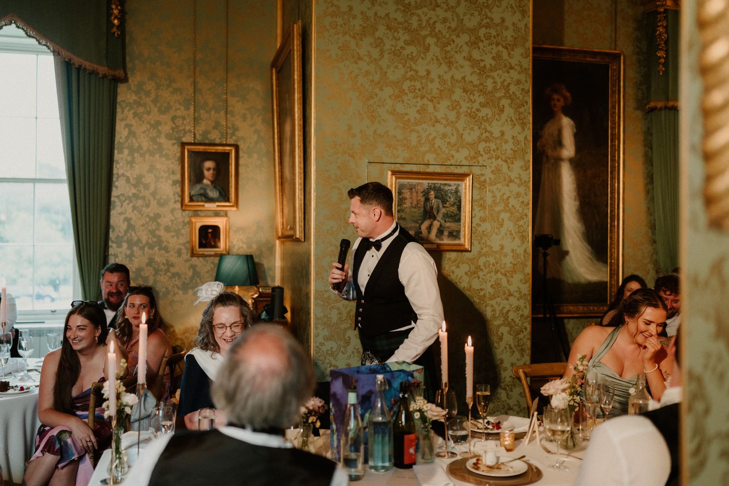 Man in a vest holds a microphone and gives a speech to seated wedding guests at candlelit tables in an elegant room with portraits.