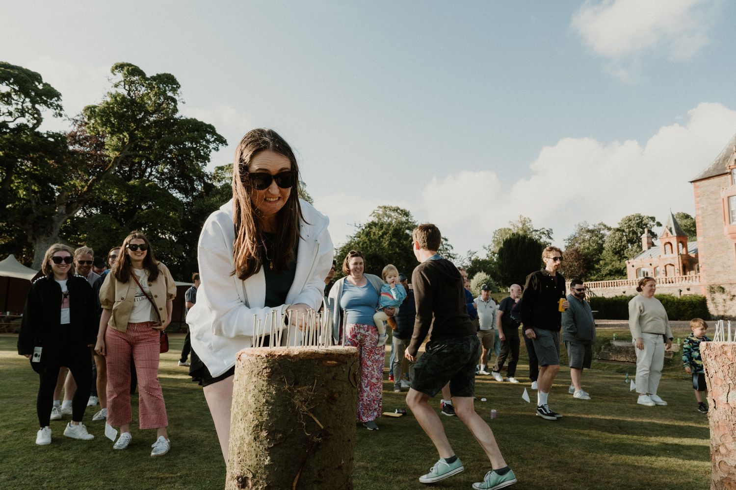 Woman in sunglasses and white jacket stands at a wooden stump while a group of people gather on a sunny park lawn behind her.
