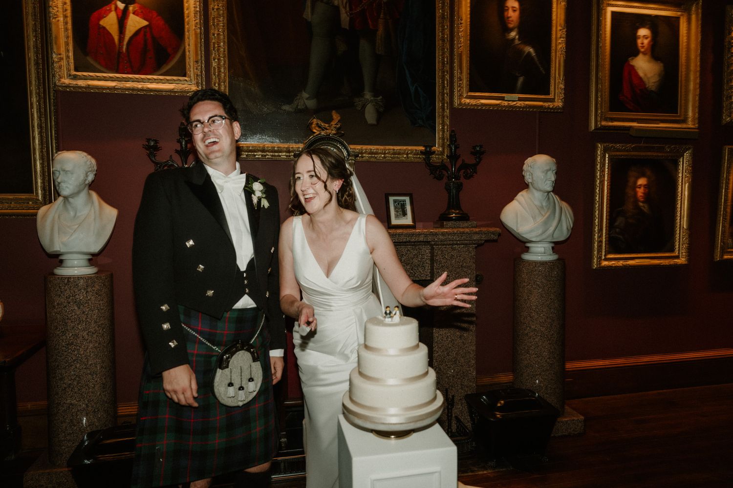 Bride in white gown and groom in Scottish kilt cut a tiered wedding cake in a gallery with portraits and marble busts.