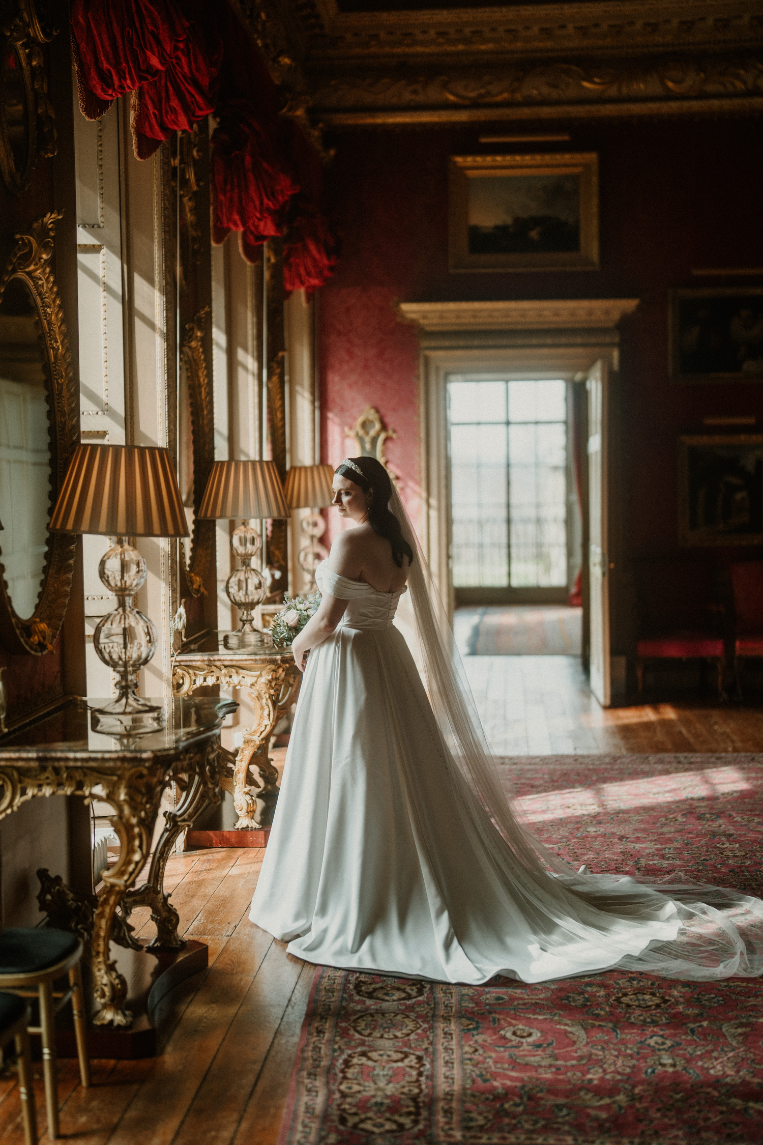 Elegant bride in white gown with veil stands in ornate golden room with red walls and hardwood floors.