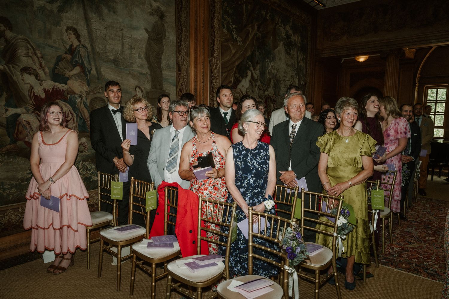 Elegantly dressed wedding guests seated in gold chairs inside an ornate room with tapestry walls.