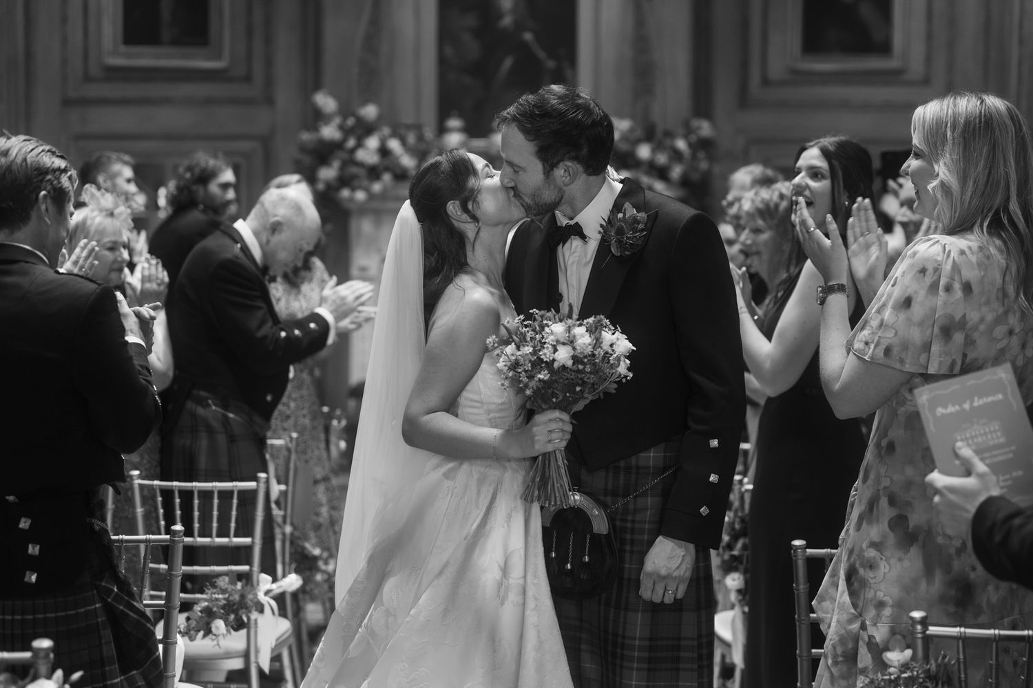 Black and white photo of a newlywed couple sharing a kiss at the altar as guests applaud in a grand venue.