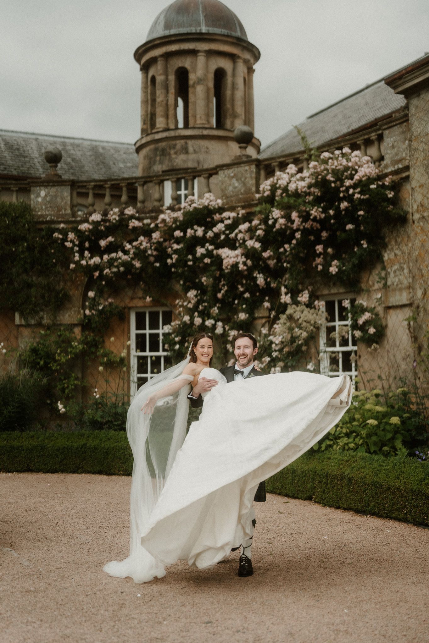 Bride and groom dancing joyfully outside a historic stone manor with climbing roses and a domed tower.
