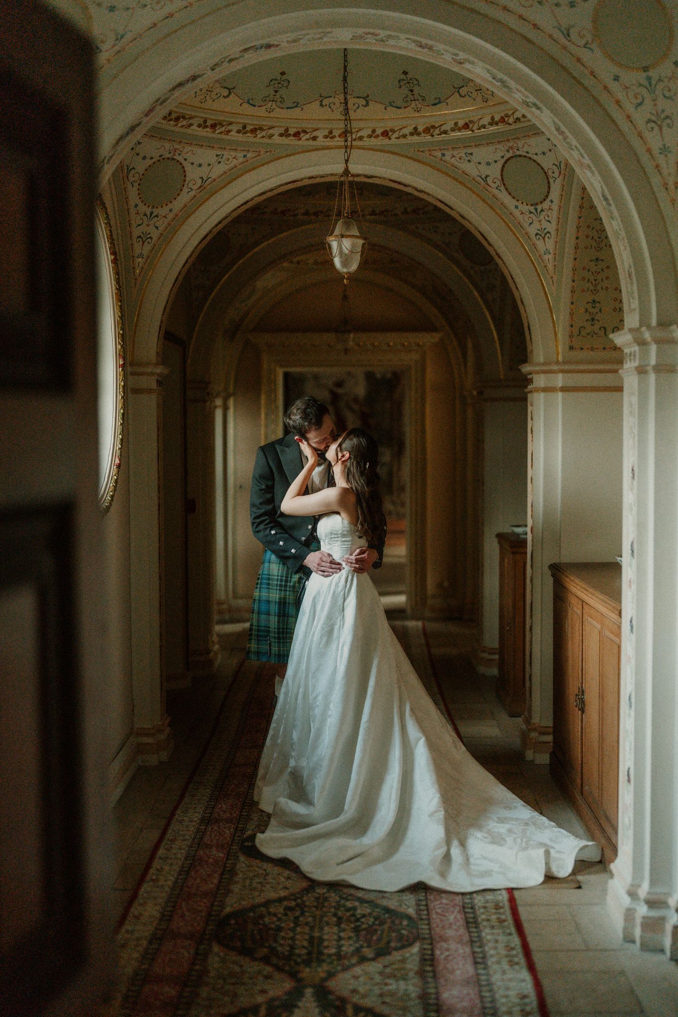 Couple shares romantic kiss under decorative arched ceiling in elegant Scottish castle corridor.