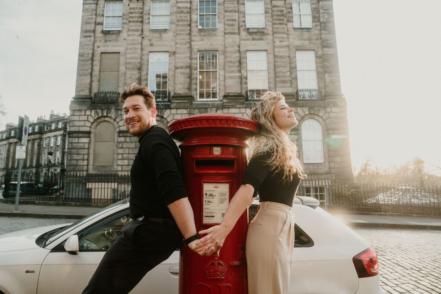 Couple holding hands leaning against a white car near a red postbox on an Edinburgh street.