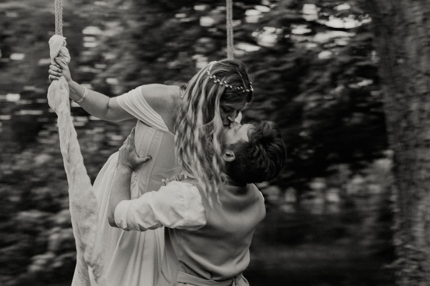 Black and white photo of a couple sharing a romantic kiss while swinging on a rope outdoors.