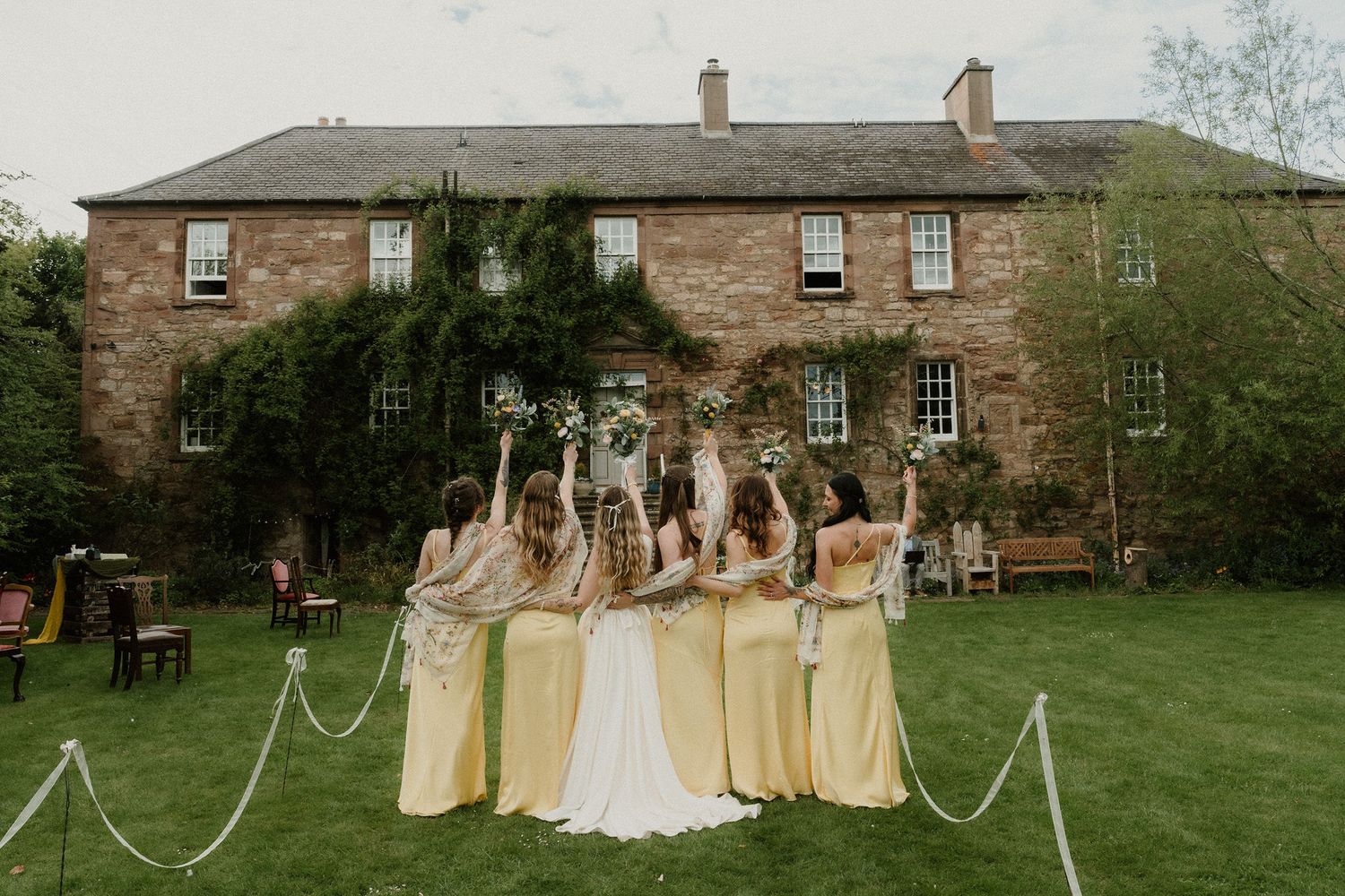 Bride and bridesmaids in yellow dresses raising bouquets outside a rustic stone manor house.