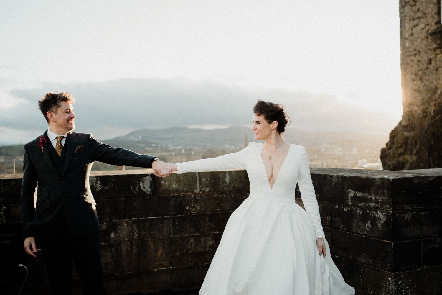 Bride and groom pose playfully on castle walls with city vista behind them.