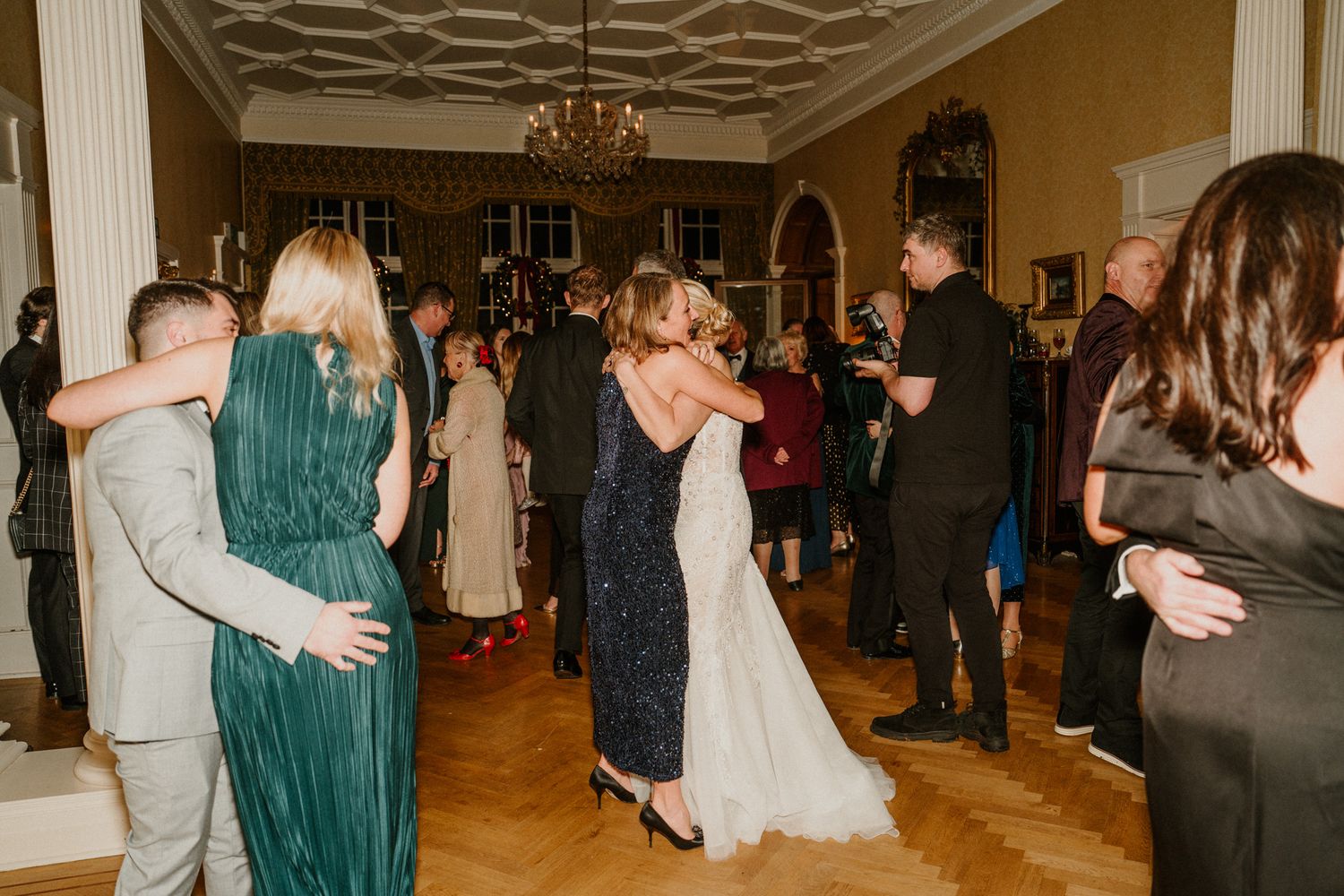 Wedding reception guests dance and mingle on a polished wooden dance floor under ornate chandeliers.