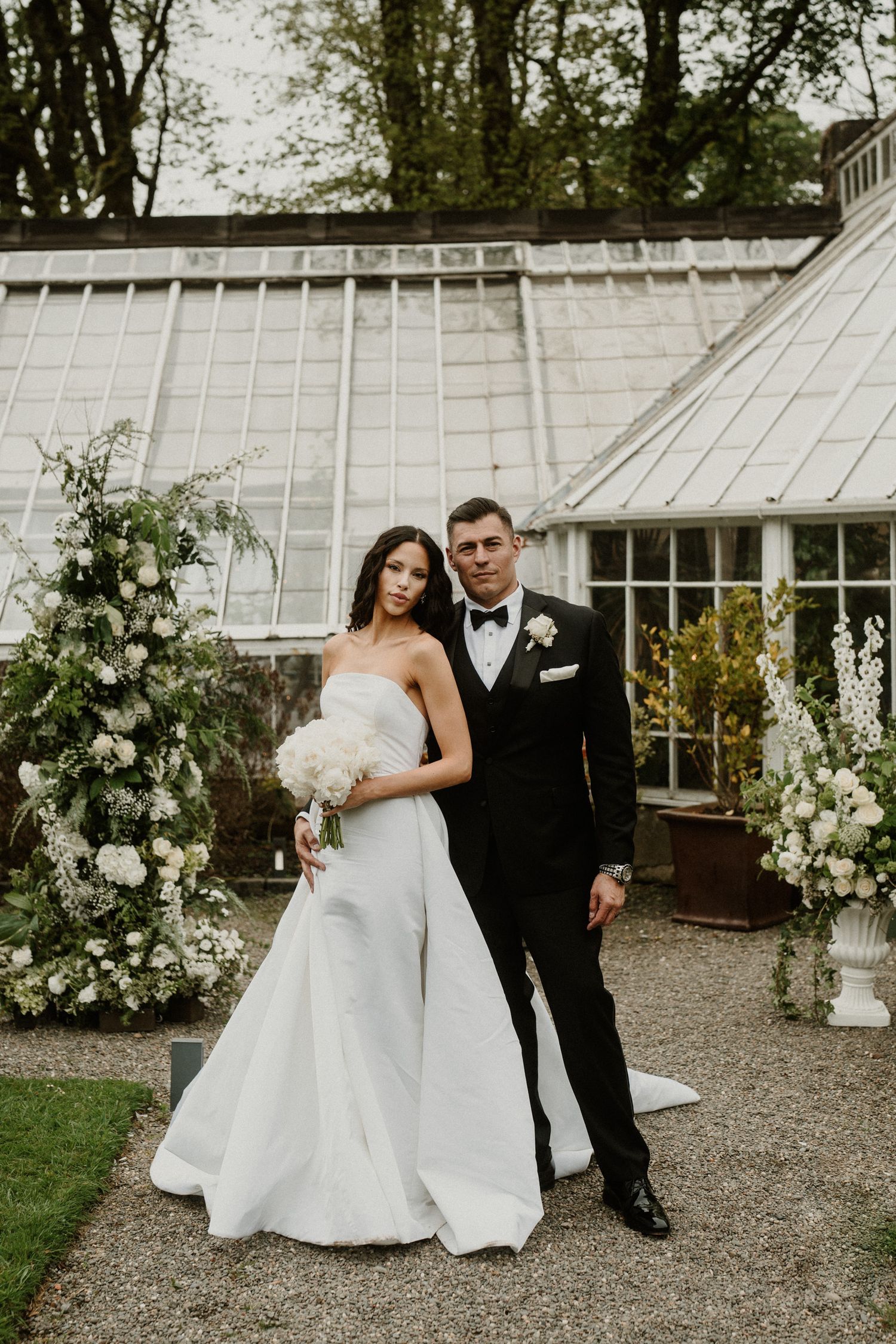 Elegant wedding couple poses in front of a greenhouse with white floral arrangements.