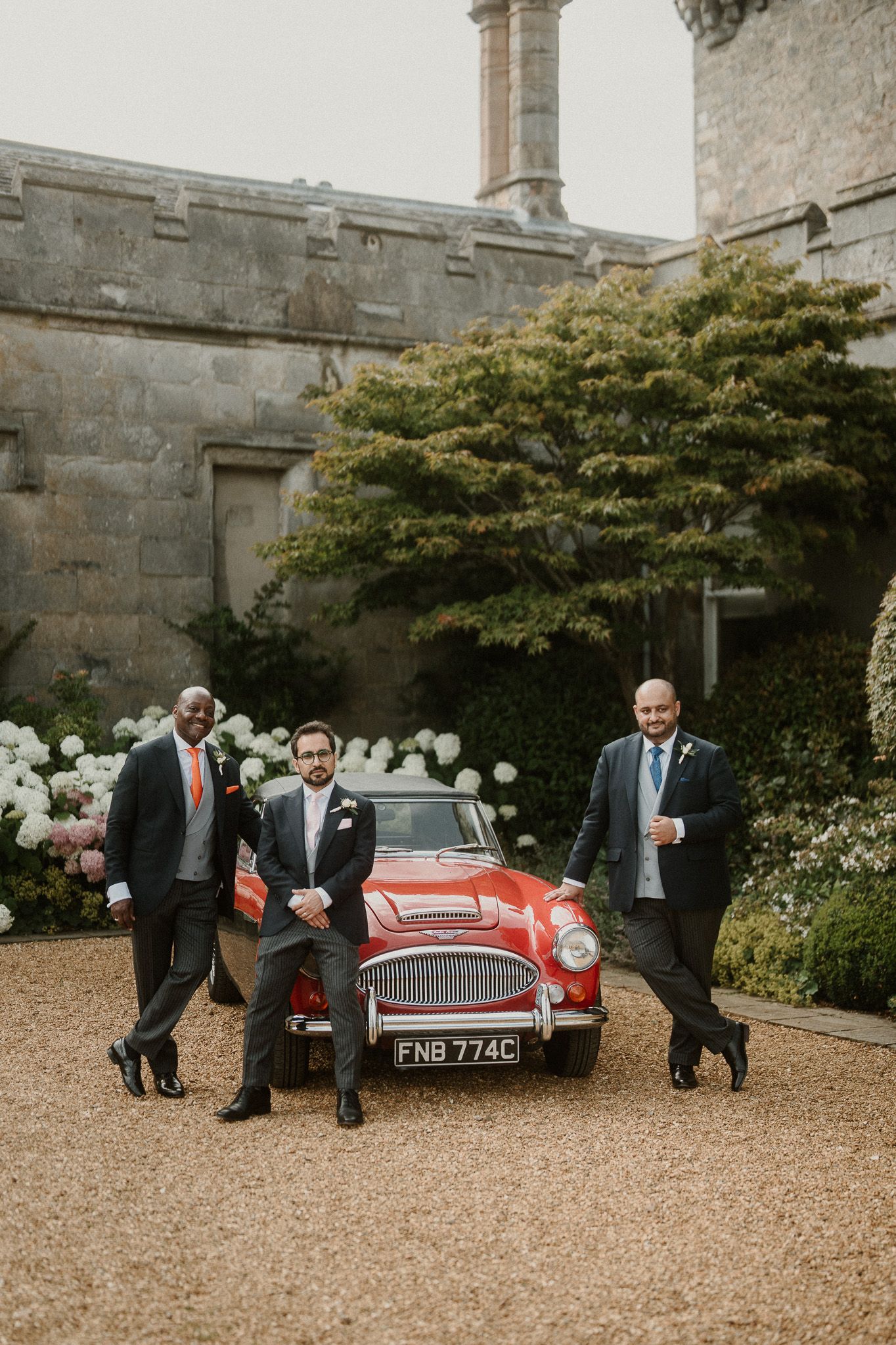 Classic red vintage car parked outside stone castle with wedding party.
