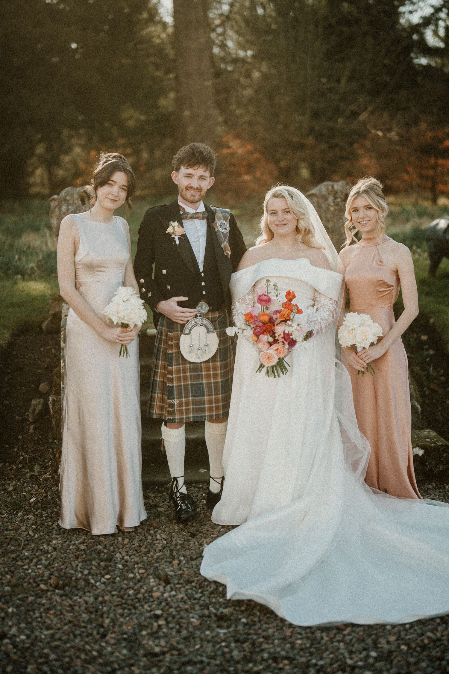 Wedding group poses in mix of formal wear and traditional Scottish attire outdoors.