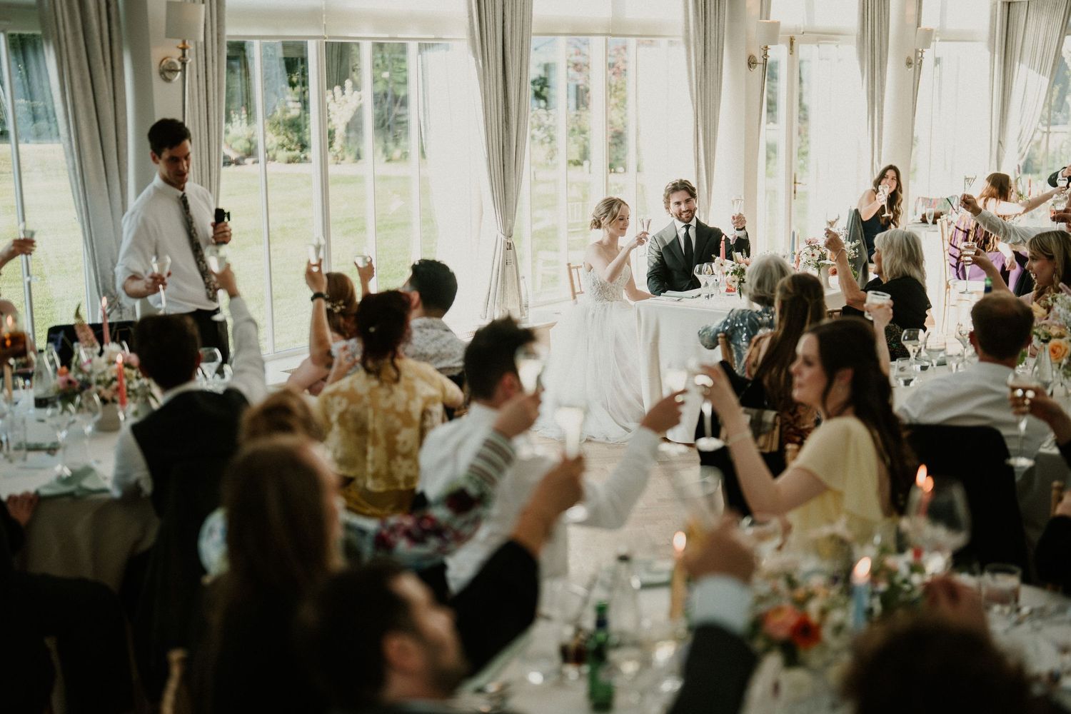 Wedding reception celebration with guests clapping in bright, window-lined venue.