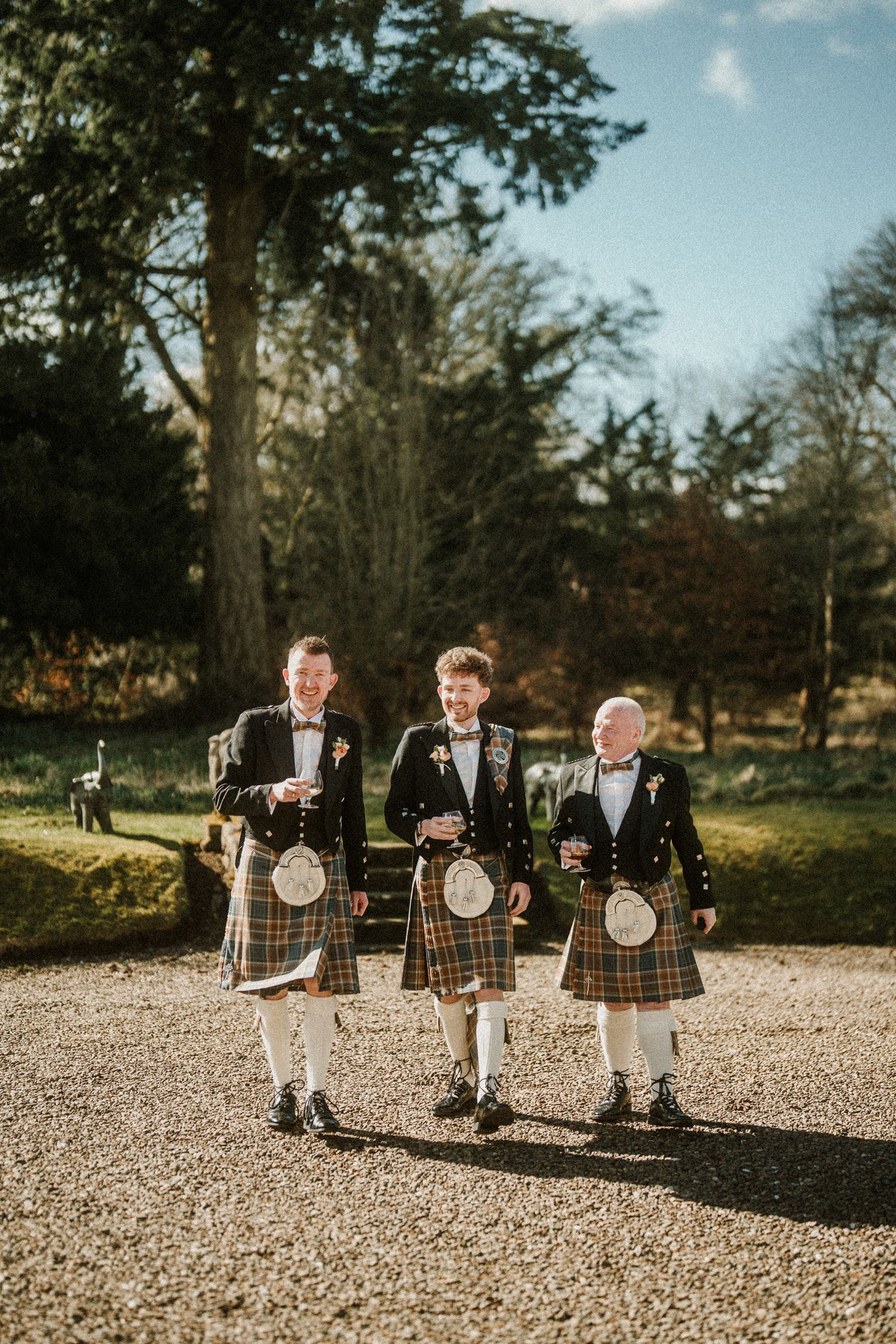 Three men in traditional Scottish kilts and formal jackets standing on a gravel path outdoors.