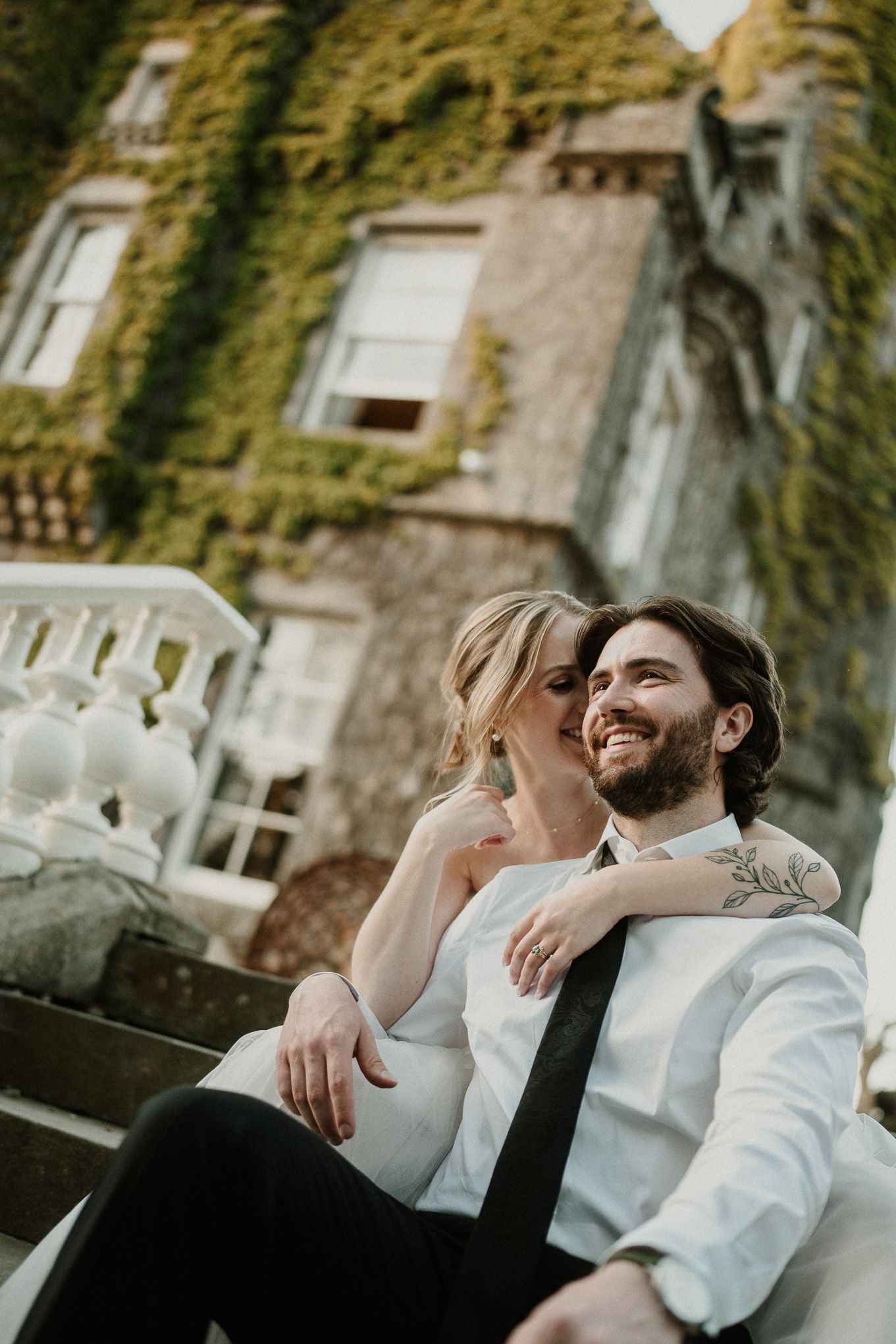 A smiling couple in wedding attire sits on steps outside an ivy-covered building, sharing a tender embrace.