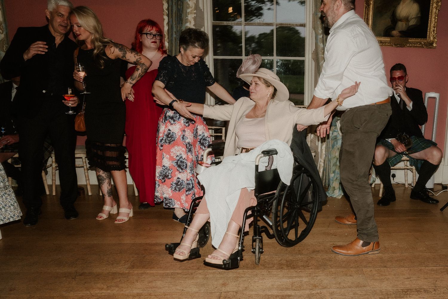 Elderly woman in wheelchair dancing with guests at a wedding reception in an elegant pink room.