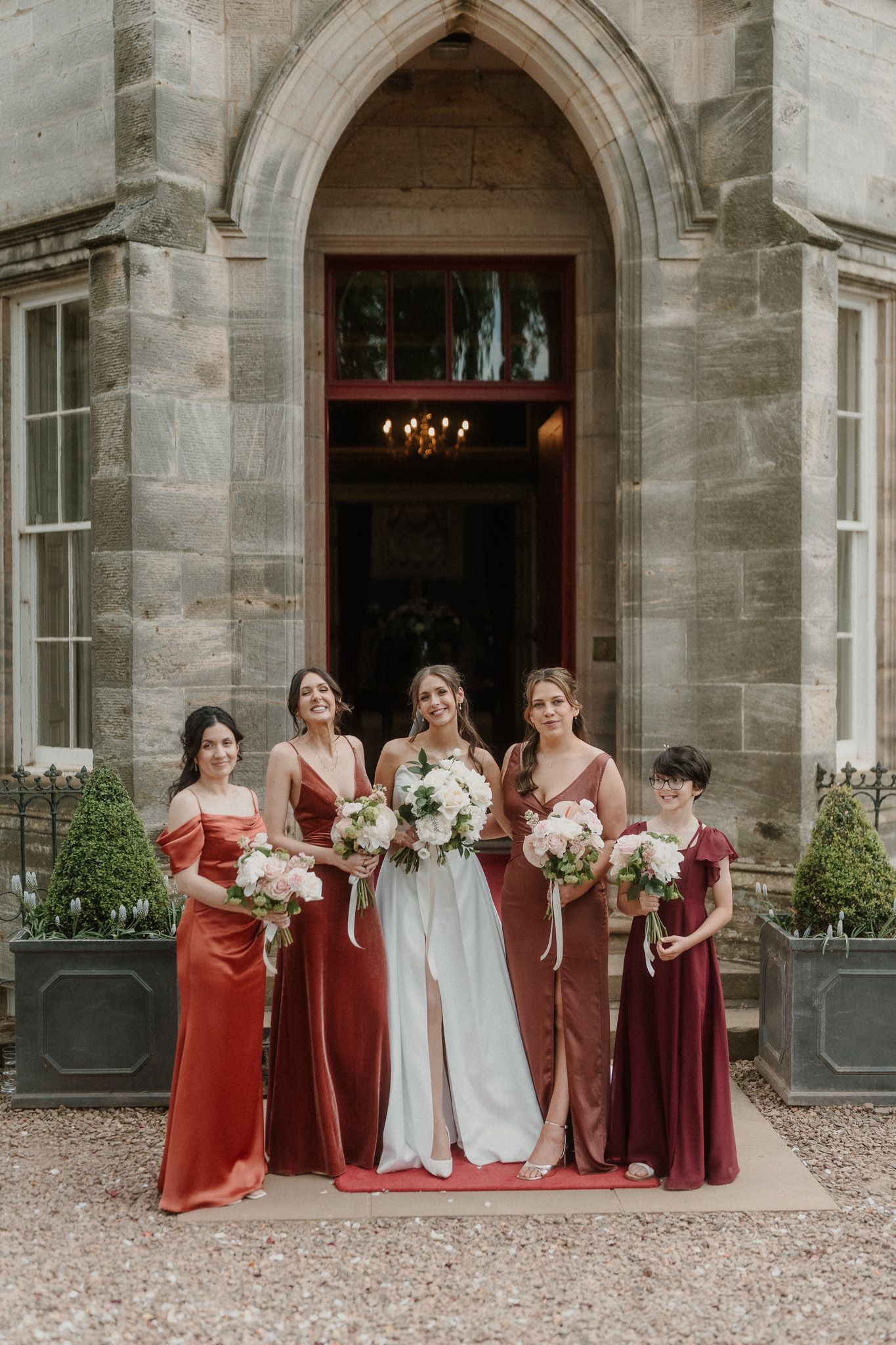 Bride in white gown poses with four bridesmaids in red satin dresses holding bouquets outside a stone manor.
