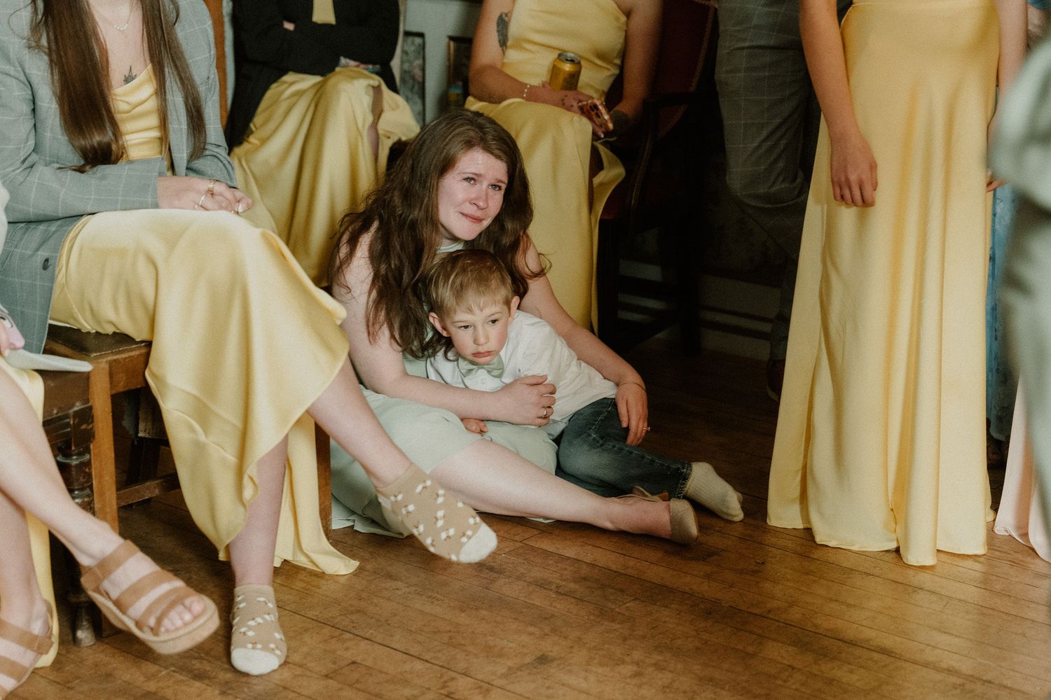 A young girl sits on a wooden floor holding a small child, surrounded by bridesmaids in yellow dresses at a wedding.