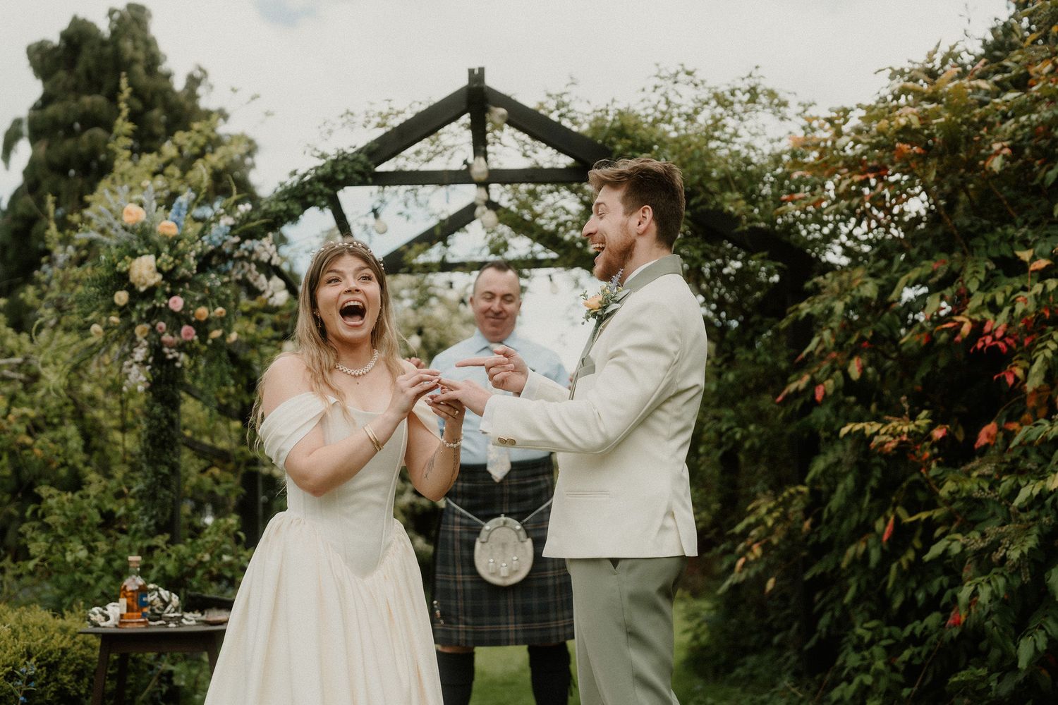 Bride laughs joyfully as groom places ring on her finger during outdoor wedding ceremony with floral arch.