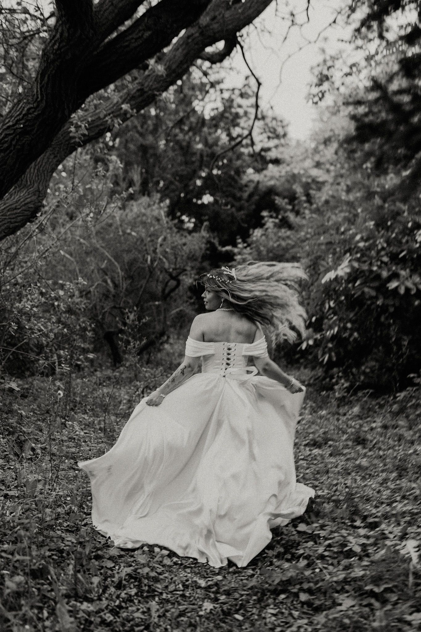 Black and white photo of bride in off-shoulder gown running through forest with hair and dress flowing.