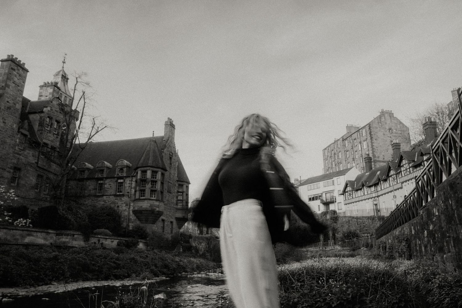 Black and white photo of a woman with flowing hair spinning outdoors near a historic stone building.