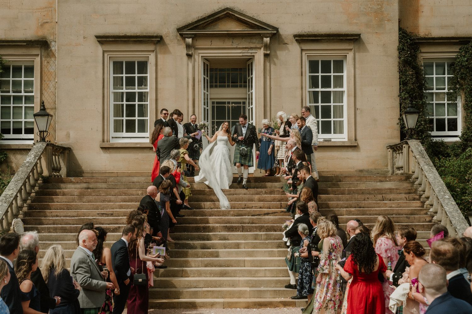 Newlyweds walk down grand stone steps surrounded by cheering wedding guests at stately venue.