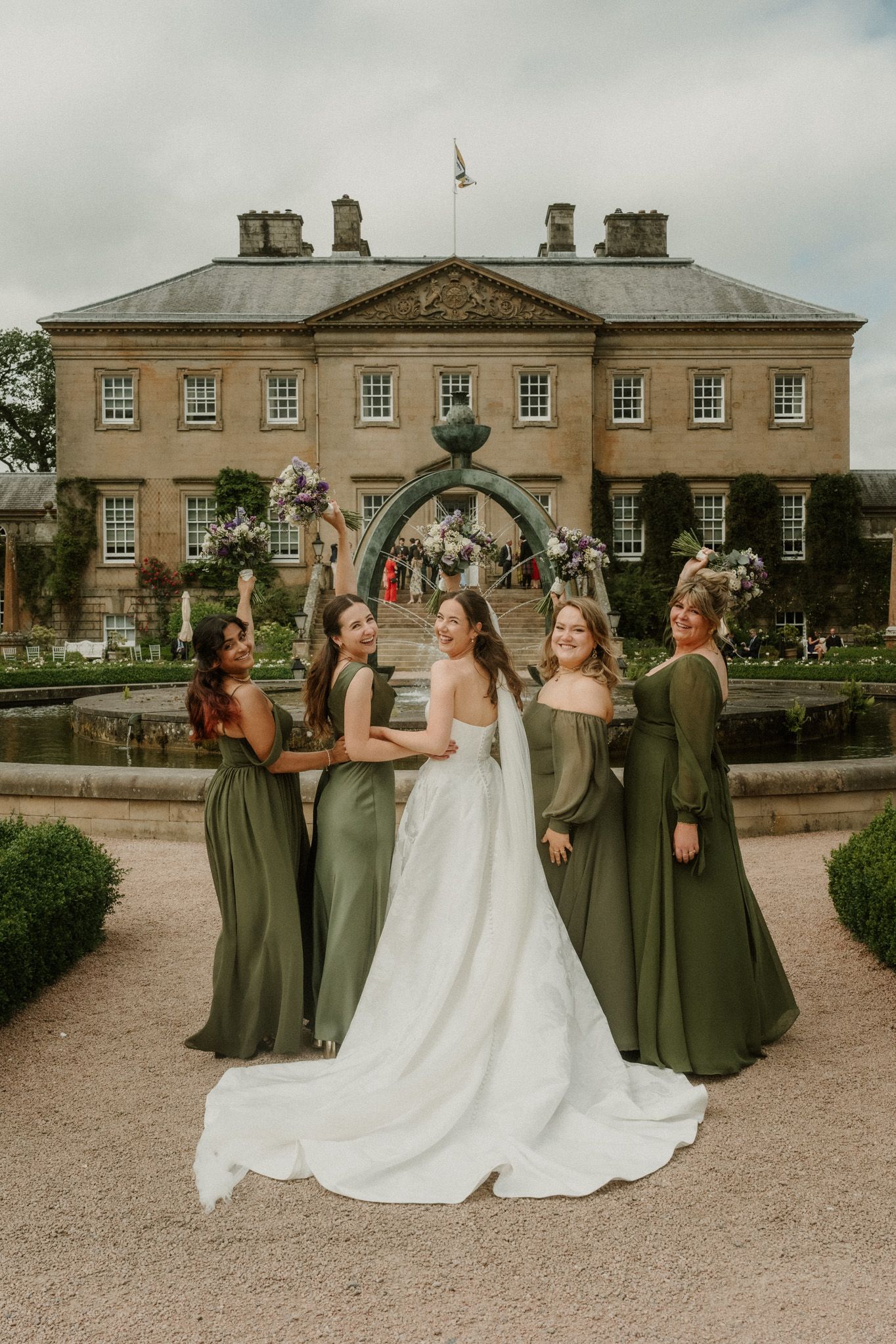 Bride in white gown poses with bridesmaids in olive green dresses outside a grand estate wedding venue.
