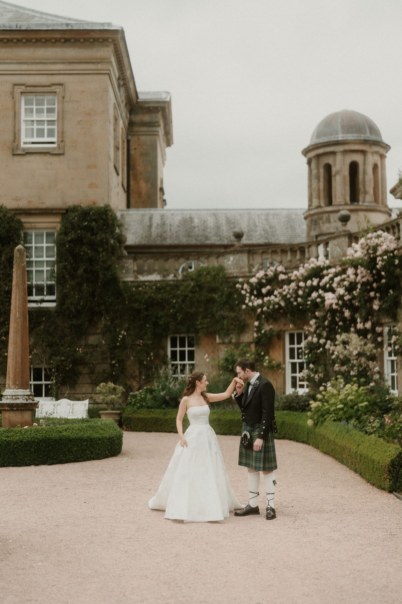 Bride in white gown and groom in Scottish kilt dance outside a grand estate with blooming gardens.