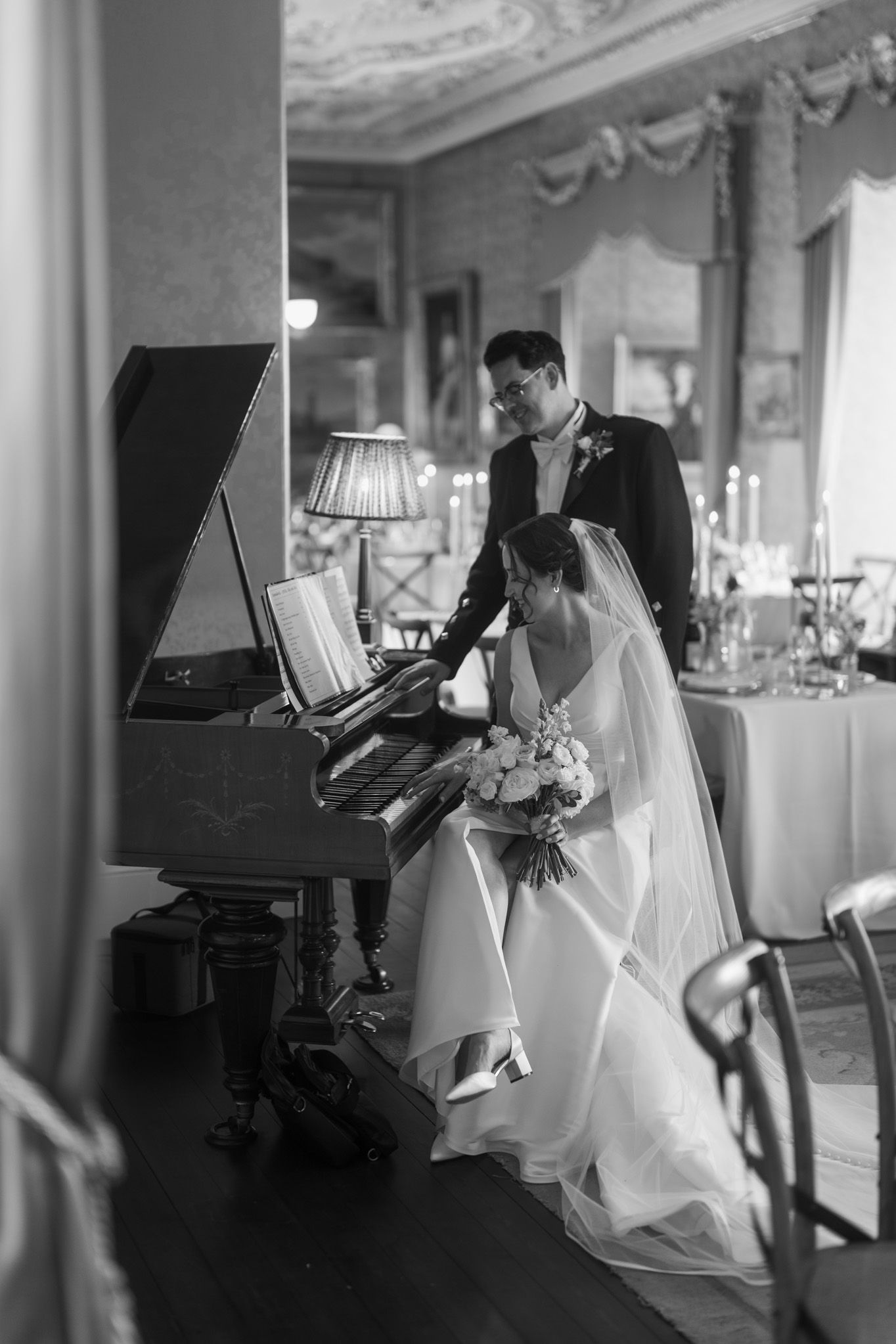 Black and white photo of a bride in a veil holding a bouquet at an upright piano while the groom stands beside her in an elegant venue.