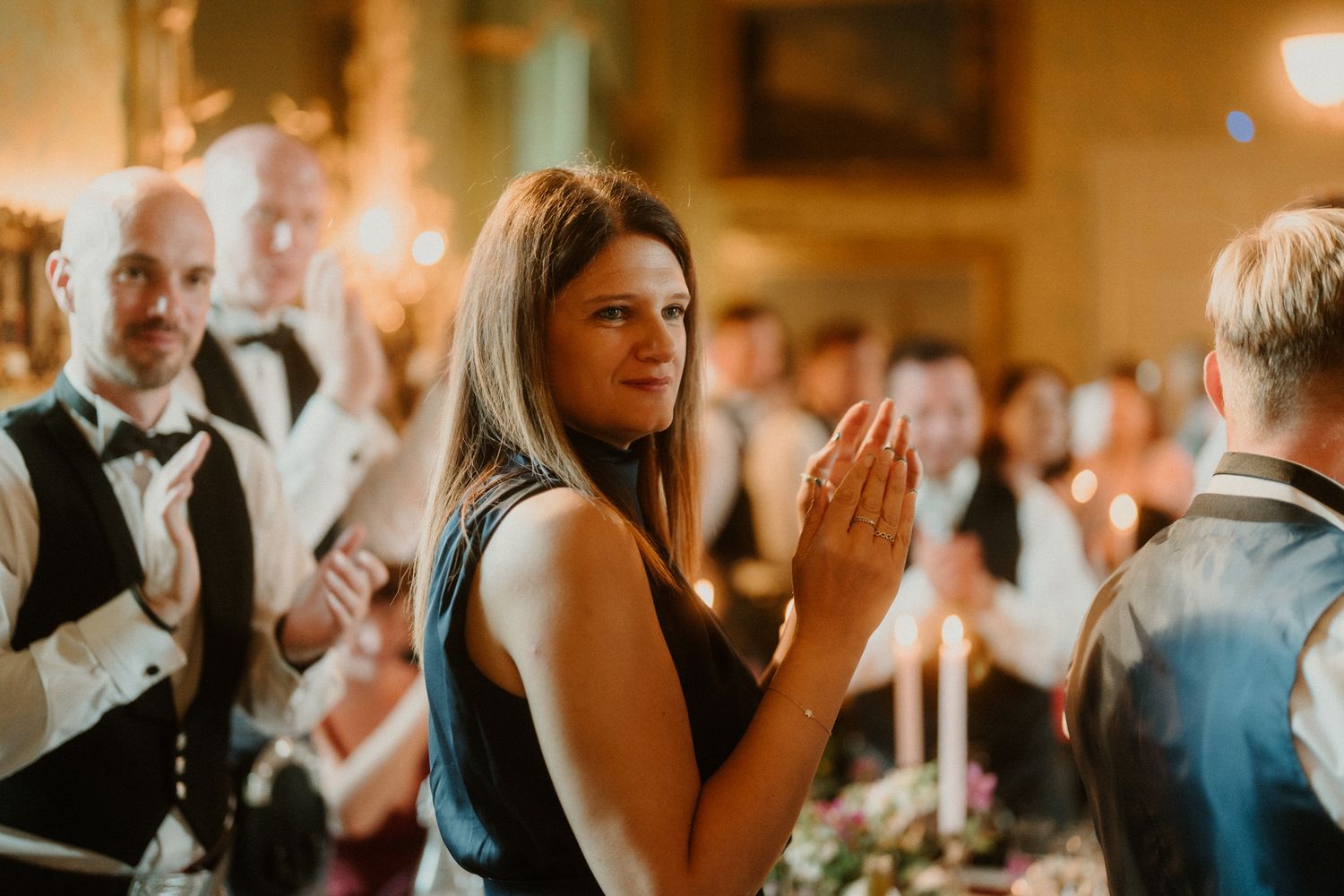 Smiling woman in a black dress applauds at an elegant candlelit banquet while formally dressed guests in vests and bow ties clap behind her.