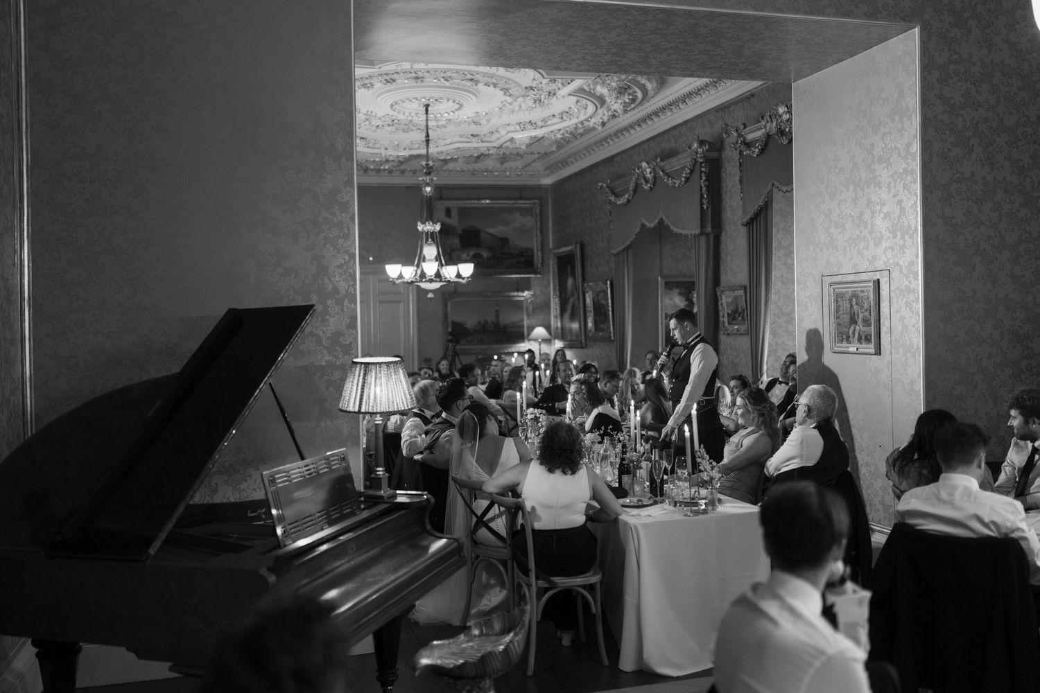 Black and white photo of guests dining at tables in an elegant room with a grand piano, chandelier and ornate ceiling.