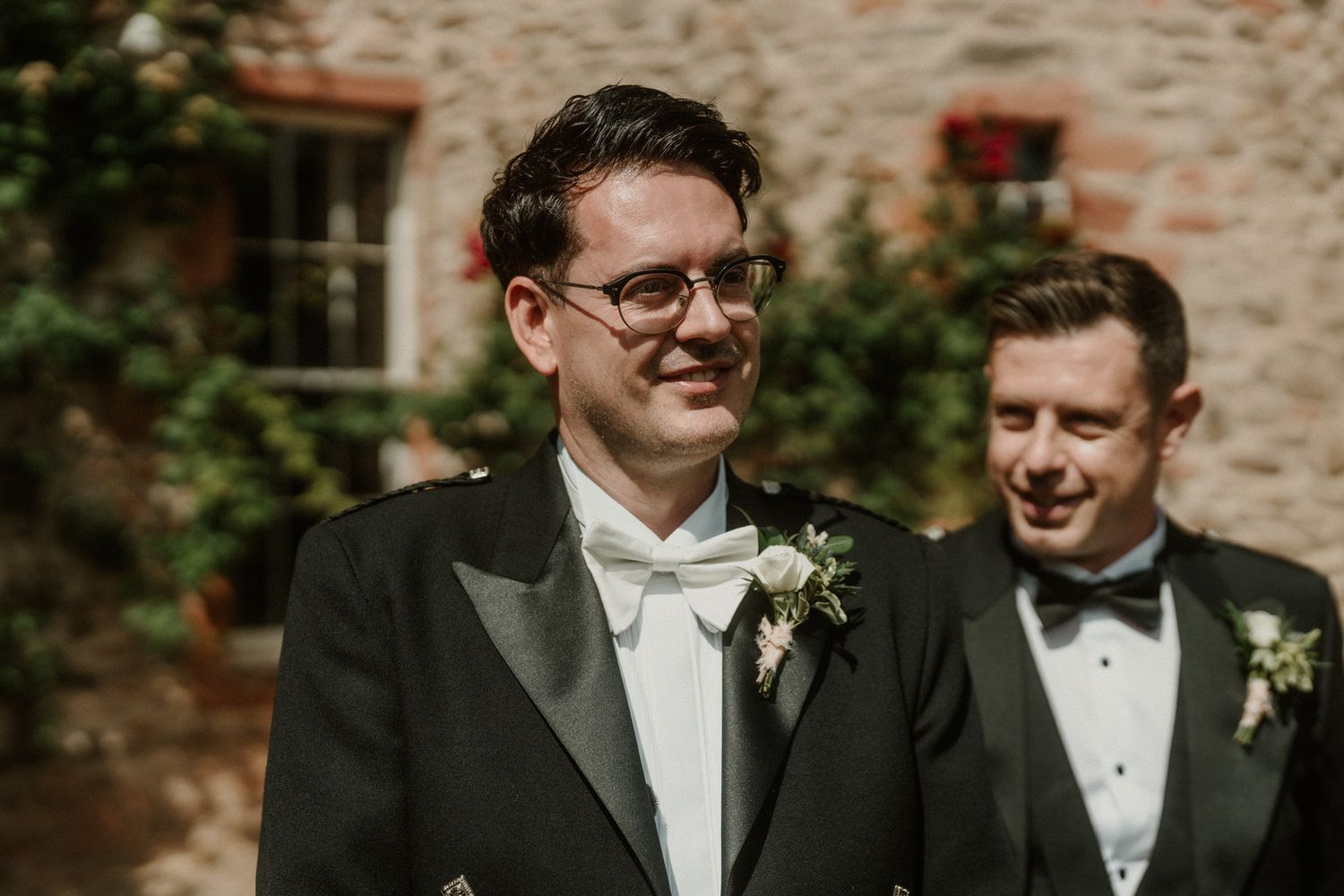 Two smiling groomsmen in black formal jackets with white floral boutonnieres stand together outside a rustic stone wedding venue.