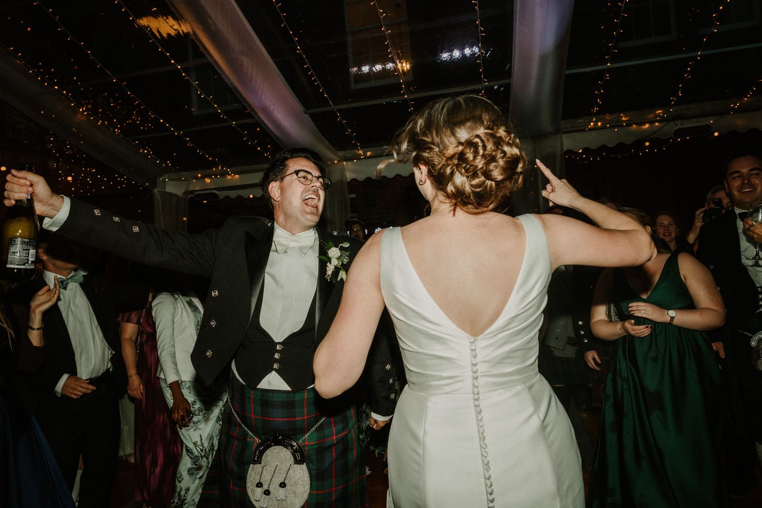 Bride in a white backless gown dances with a smiling groom in a Scottish kilt and tweed waistcoat at a lively evening wedding reception.