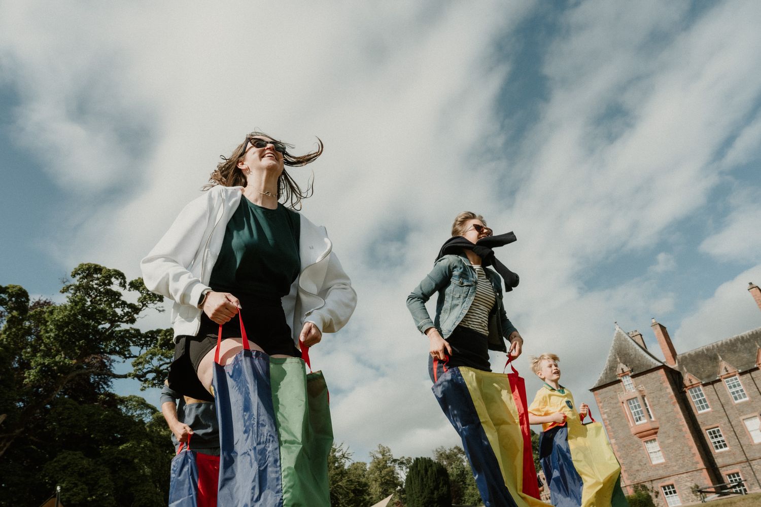 Two women in casual jackets jump joyfully in a sack race on a grassy lawn beneath a bright cloudy sky, with a stone building behind.