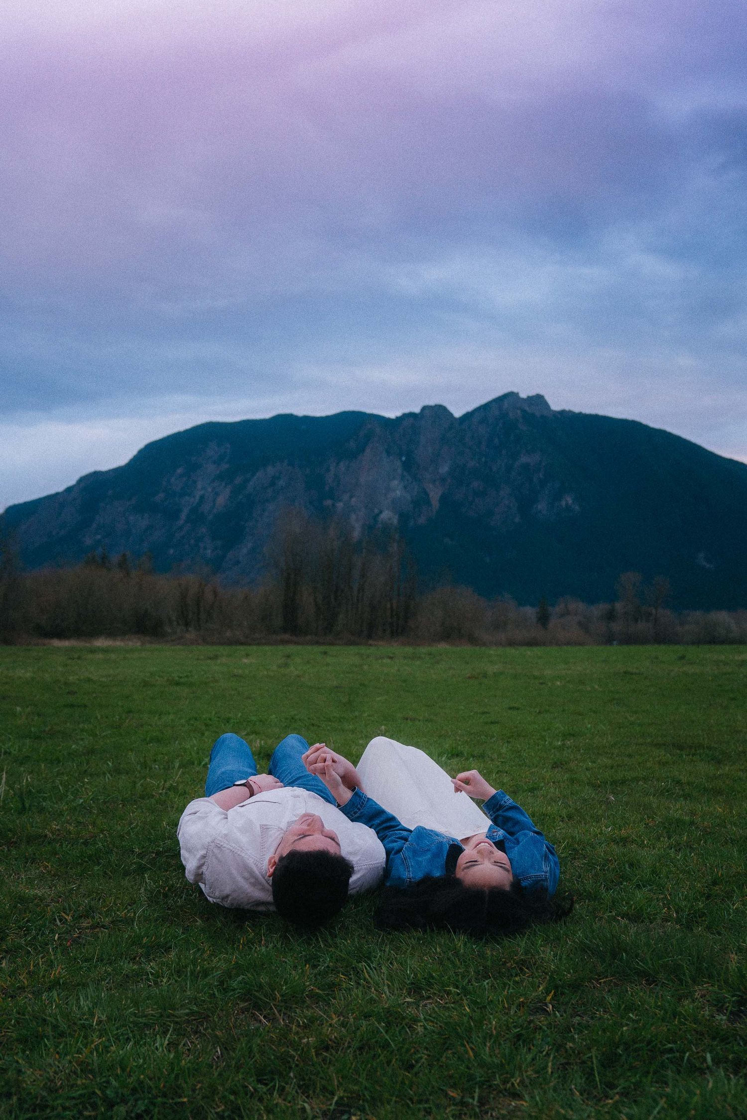 Couple lying on green grass field with mountain backdrop under dramatic purple cloudy sky.