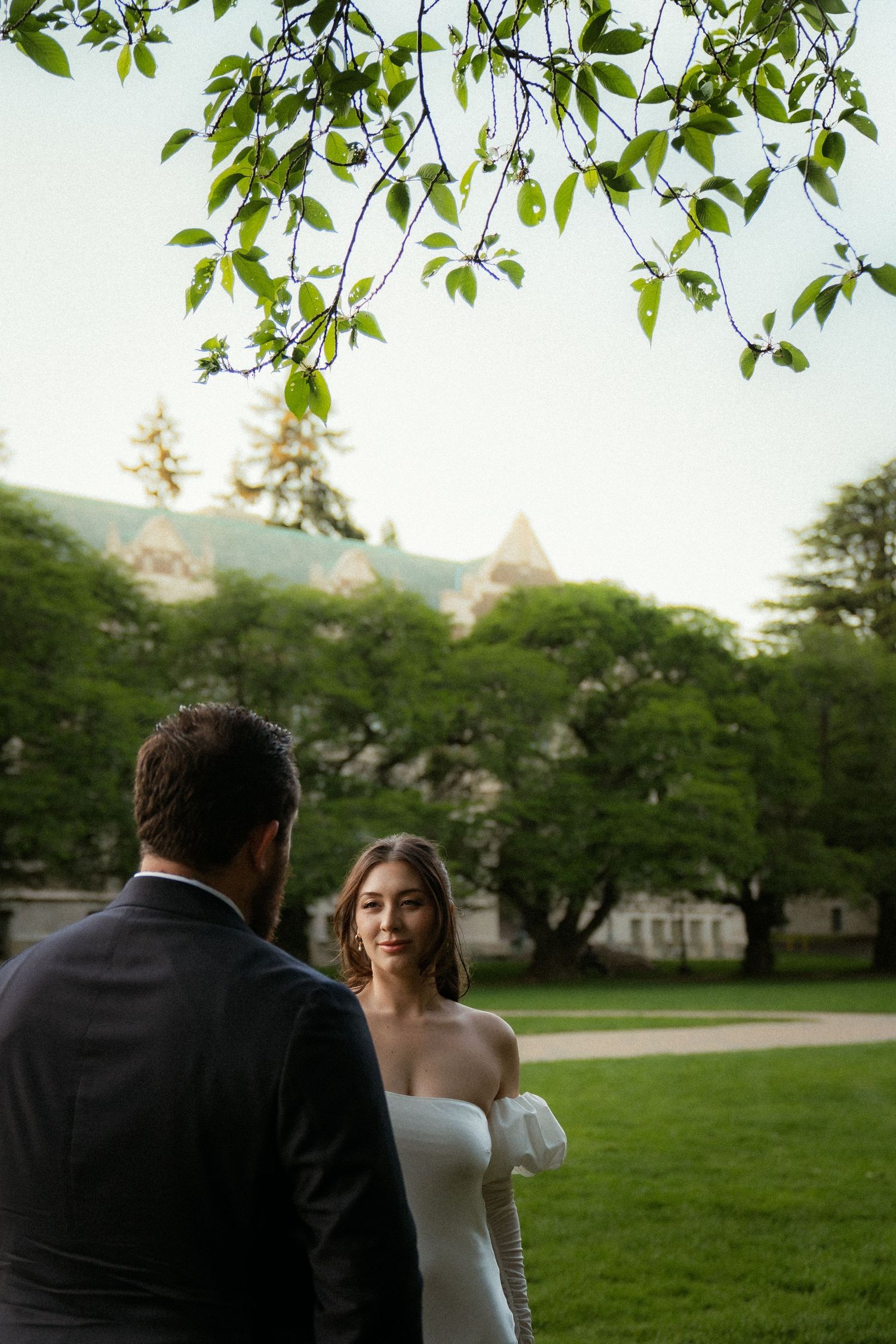 Bride in white off-shoulder gown smiles at groom in suit on lush green lawn with historic building backdrop.