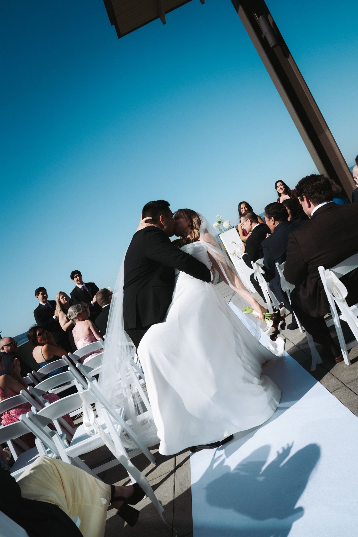 Bride and groom share a kiss at an outdoor wedding ceremony under a clear blue sky with guests seated in white chairs.