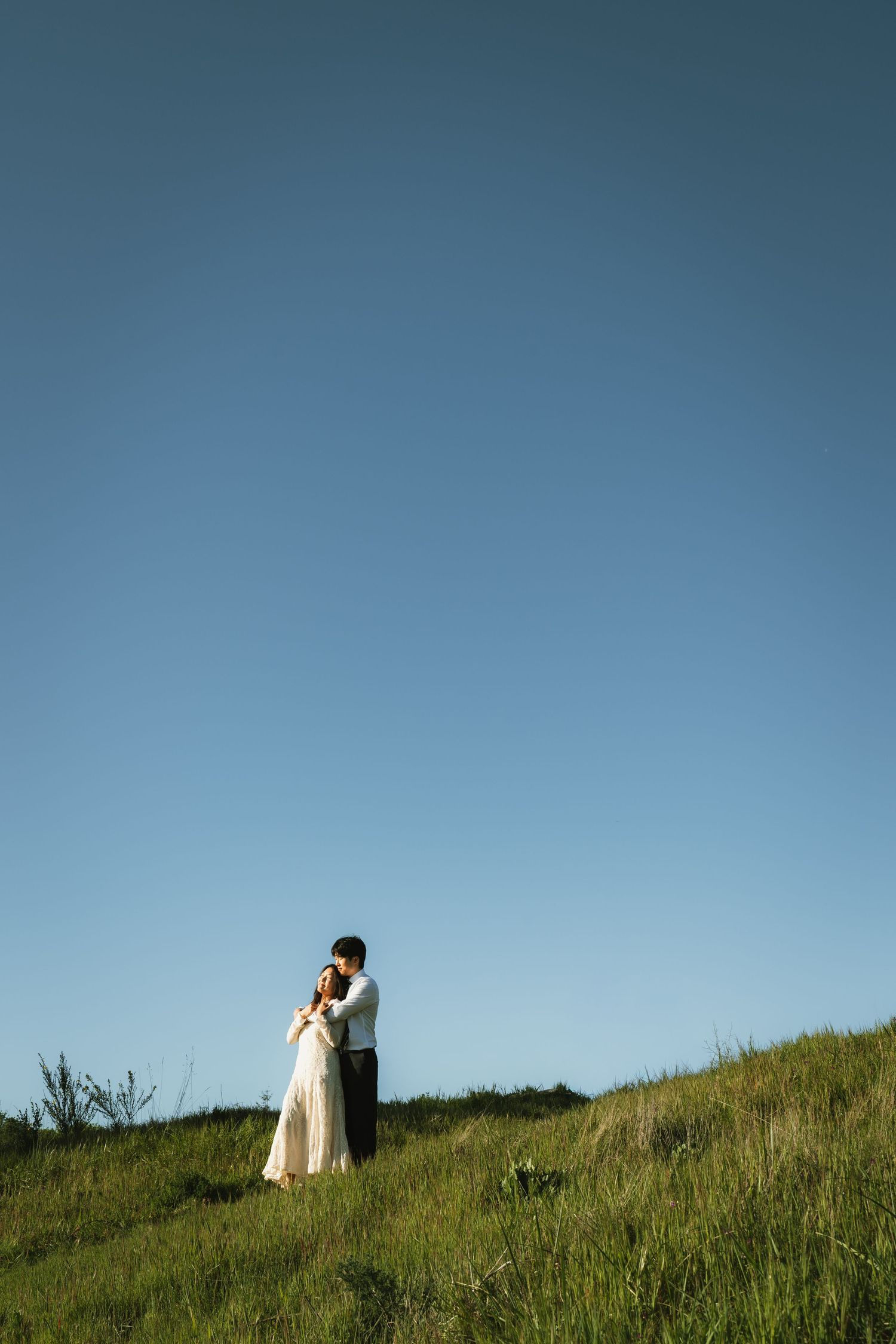 Couple embracing on a green hillside beneath a clear blue sky in a romantic outdoor portrait.